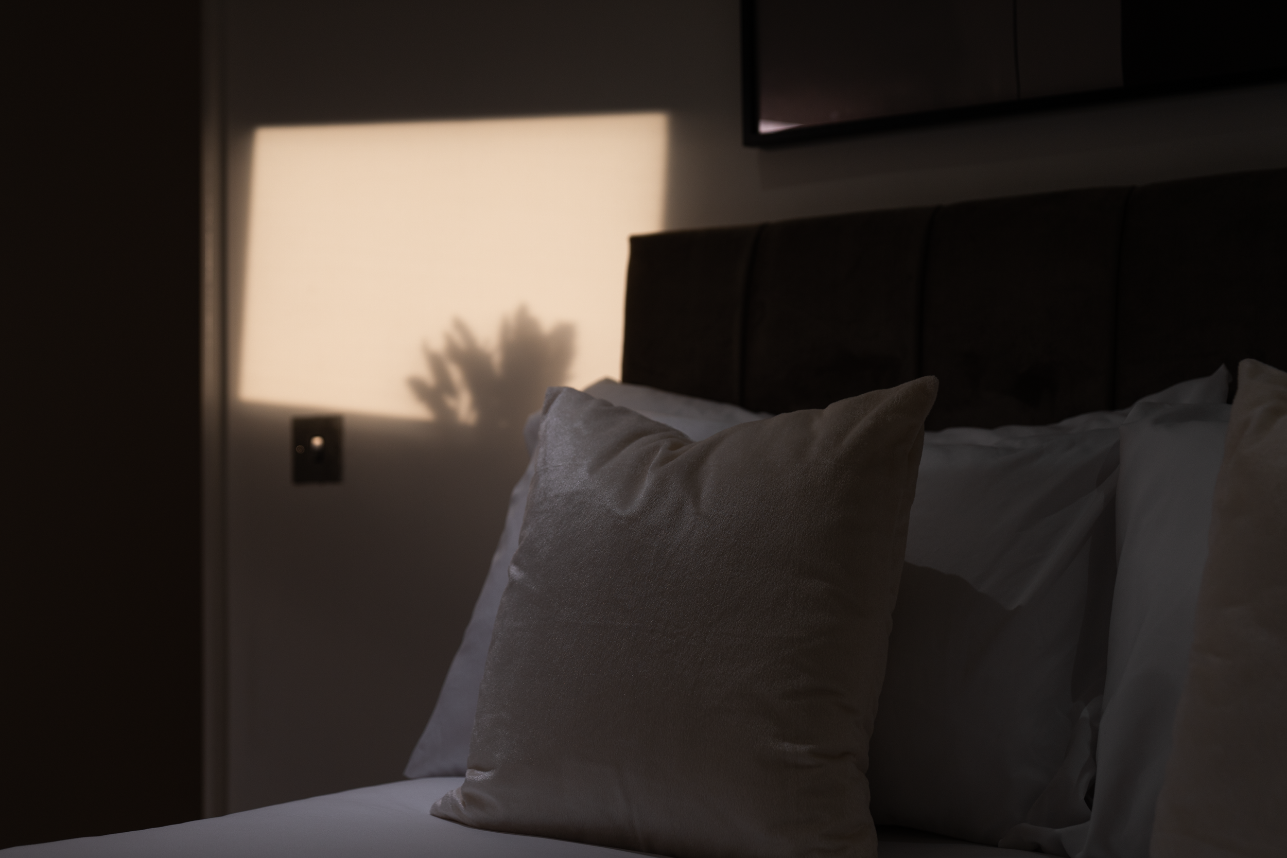 A neatly made bed with pillows in a dimly lit room, sunlight casting shadows through a window.