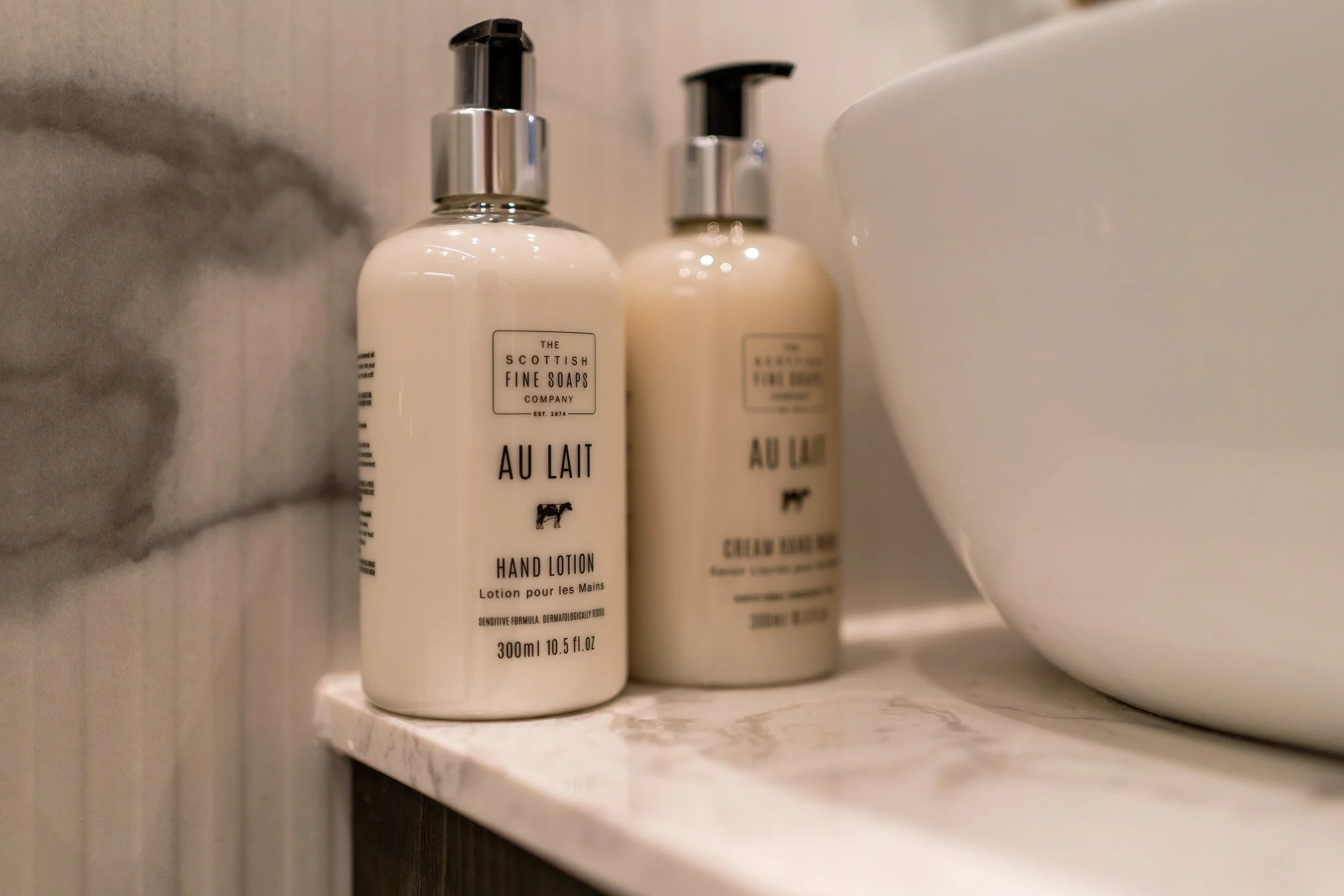 Two bottles of hand lotion and one bottle of cream soap on a marble surface next to a white bowl in a bathroom.