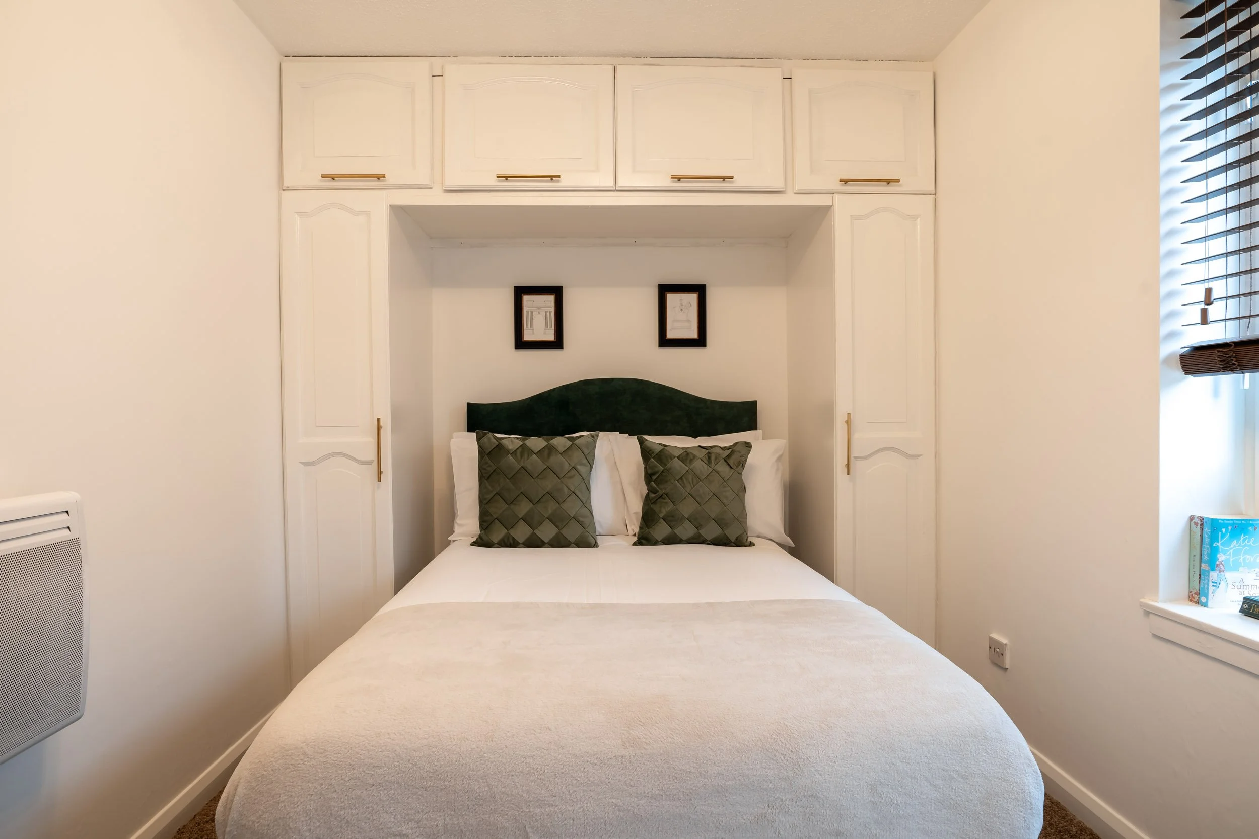 A neatly made bed with white sheets, green velvet headboard, two decorative green pillows, and two white pillows, in a room with white walls and built-in white cabinets above the bed.