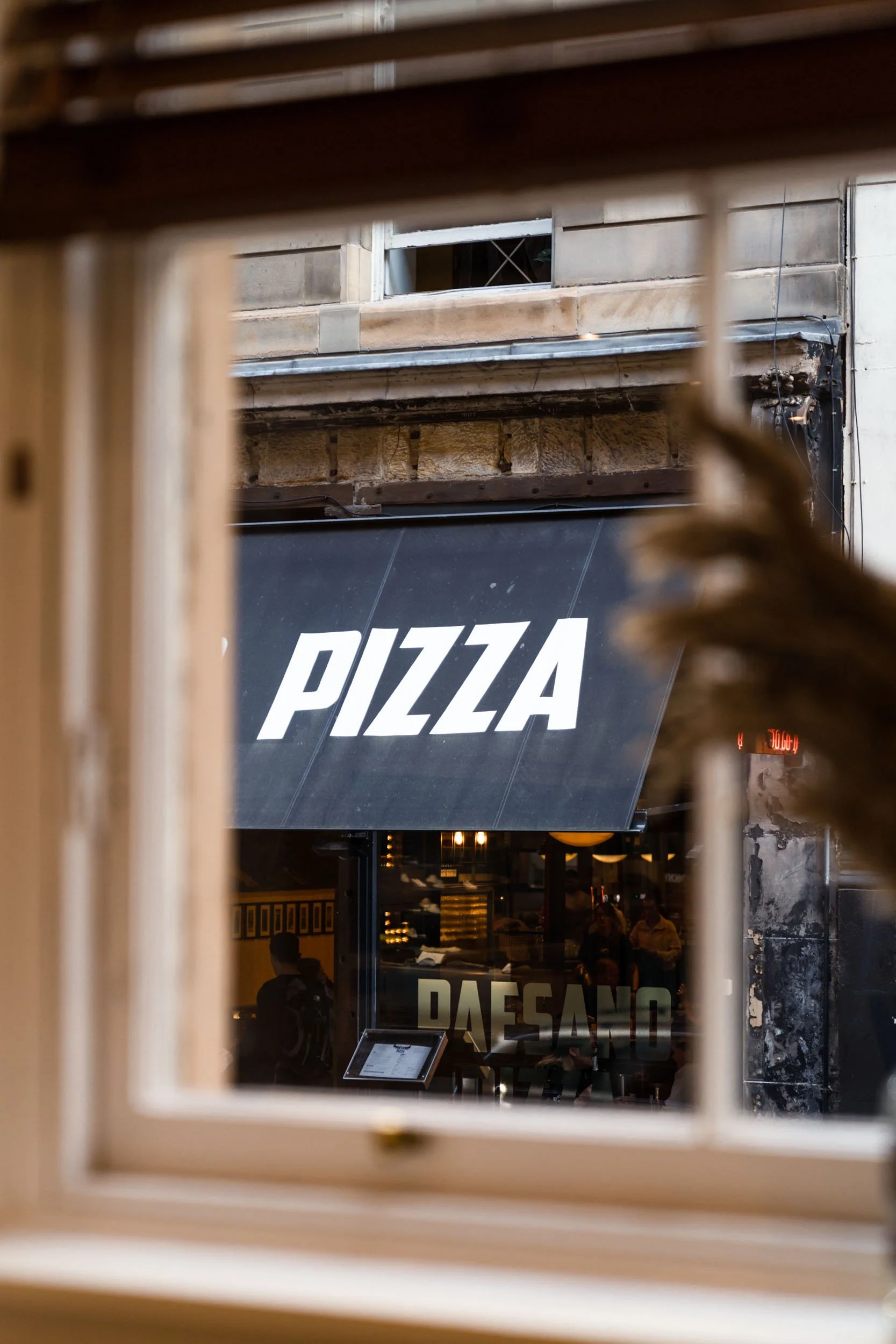 View of a pizza restaurant sign seen through a window frame, with the word 'PIZZA' on a black awning outside.