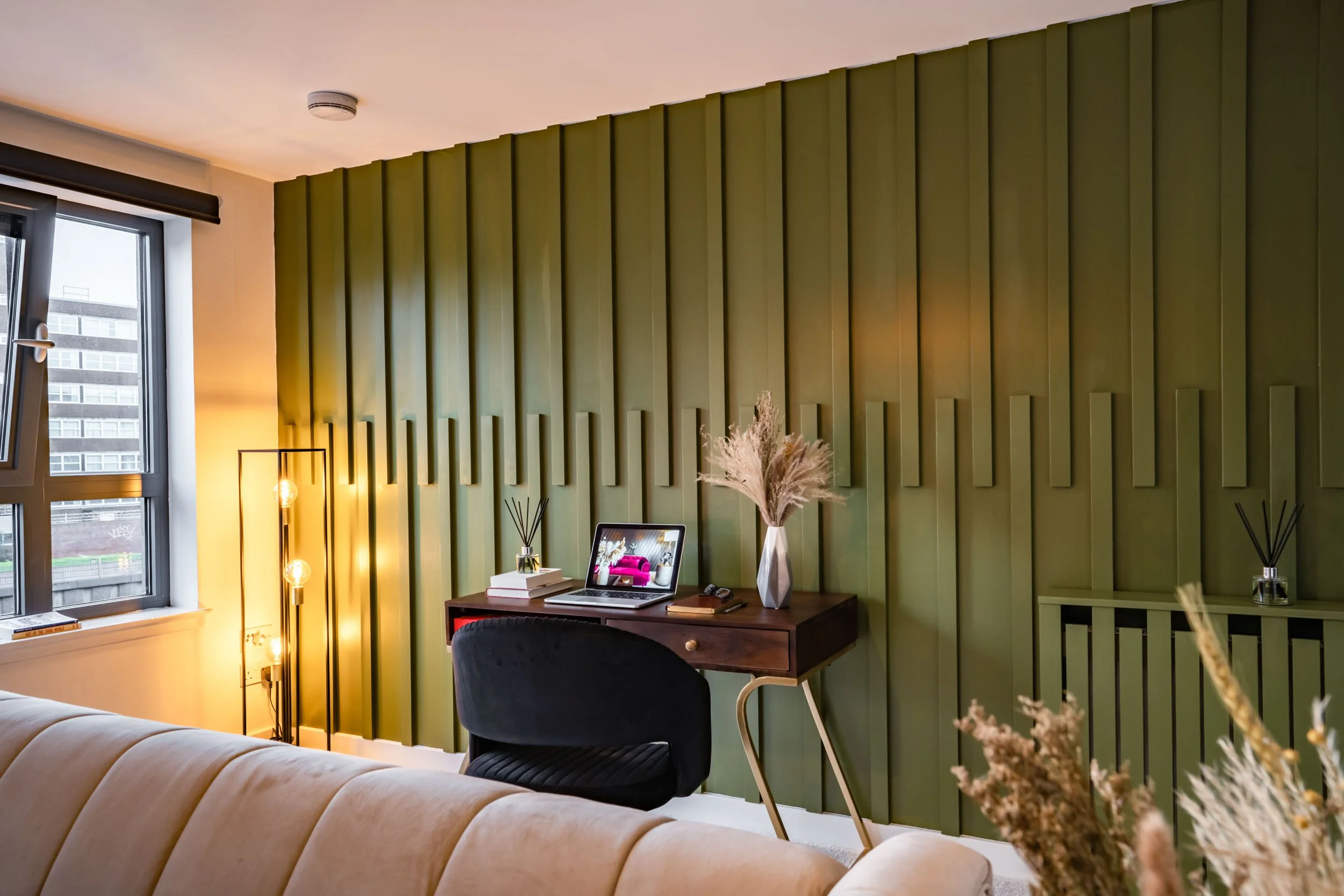 A modern living room corner with a beige couch, a wooden desk with a black chair, a laptop, books, dried flowers, and a floor lamp, set against a green textured accent wall with tall windows nearby.