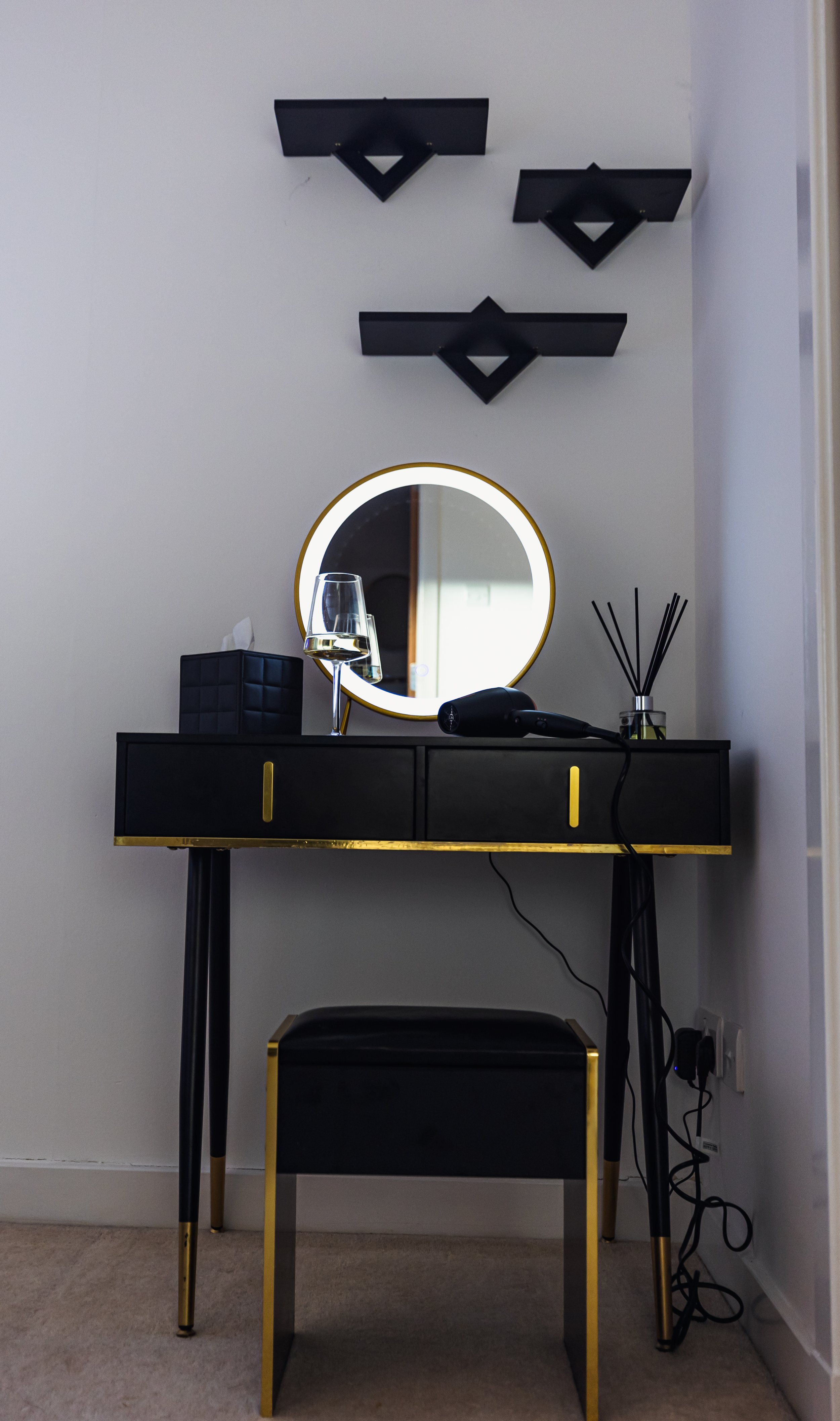 A modern black and gold vanity table with a round mirror, a wine glass, a diffuser, a hairdryer, and a black decorative box, against a white wall with black geometric wall shelves.