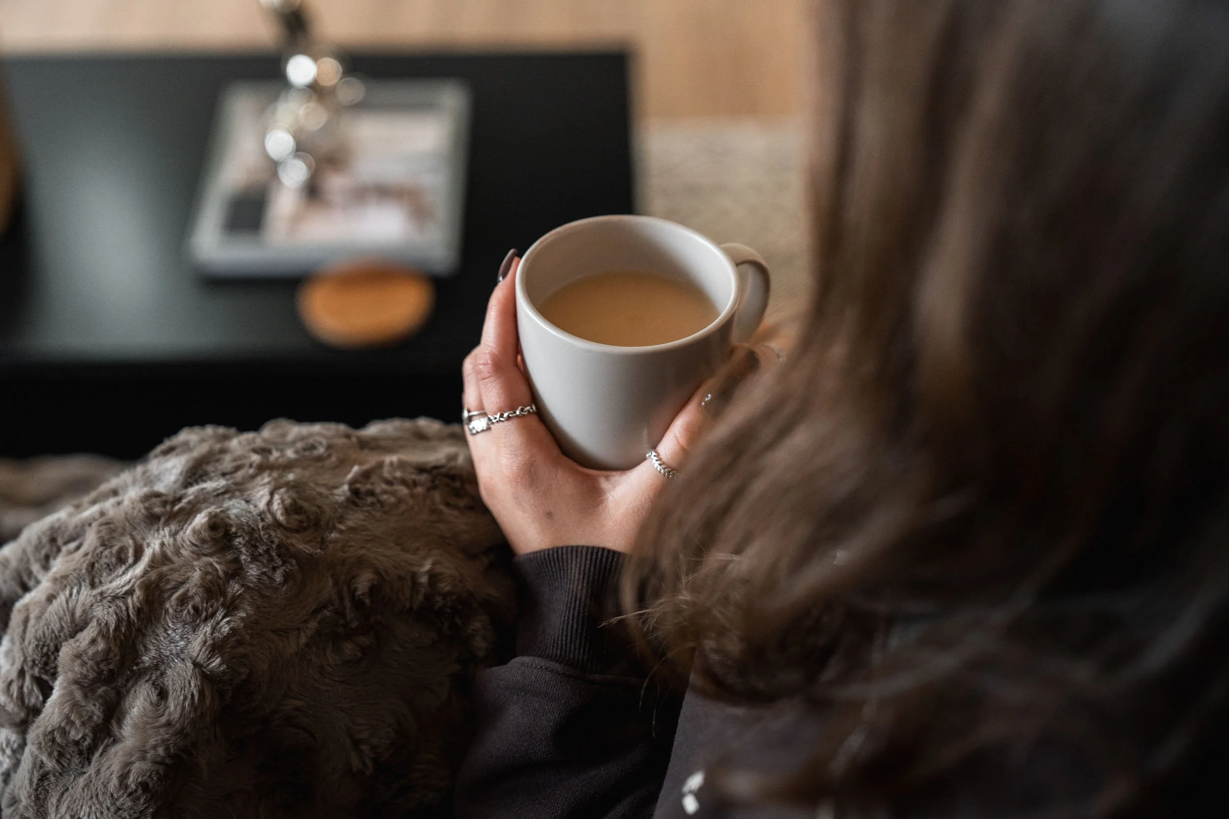 Person holding a white mug filled with coffee, sitting on a couch with a textured blanket. In the background, a black table has a framed photo and a cookie.