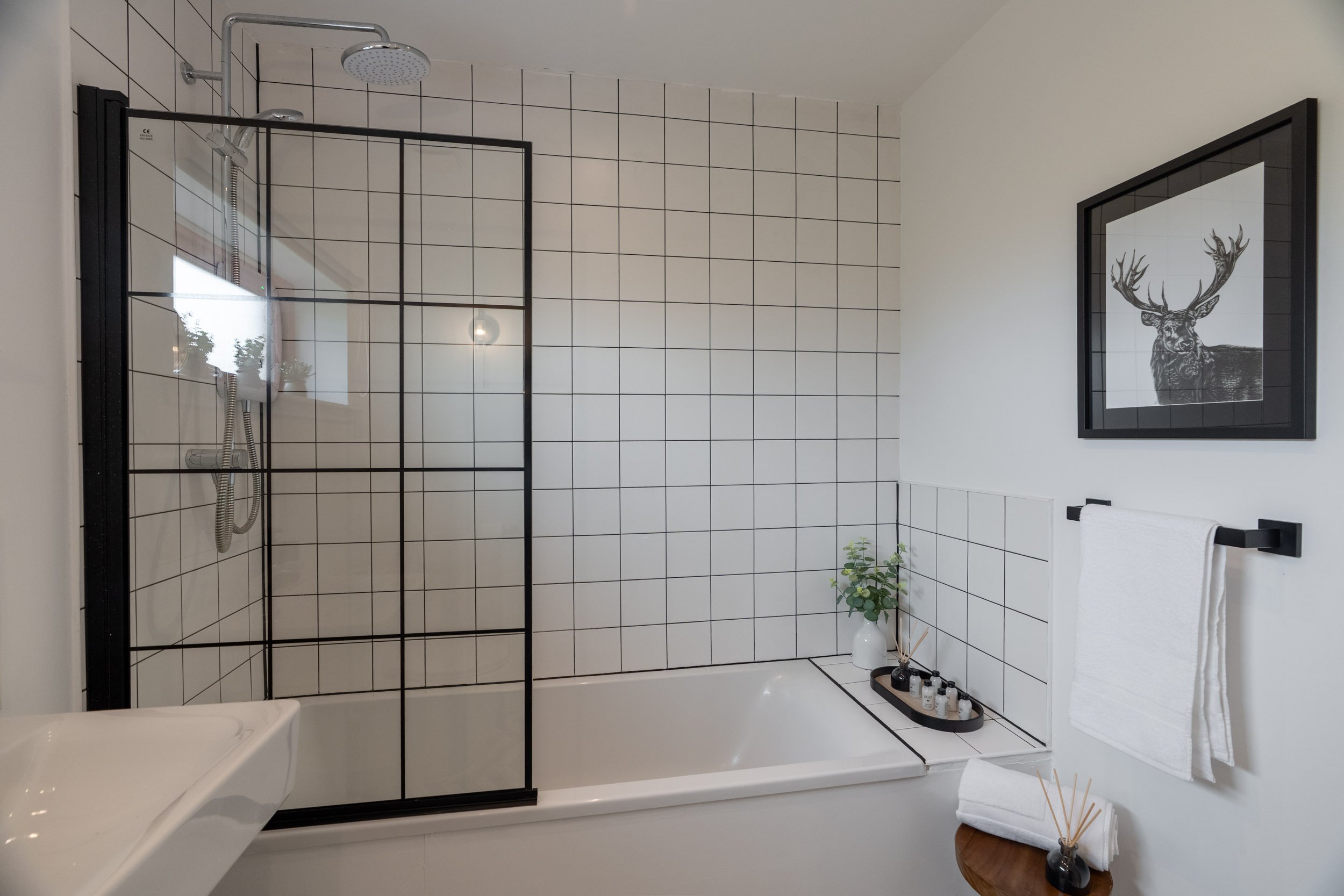 Modern bathroom with white tiled walls, a built-in bathtub, a glass shower enclosure with black frame, a framed black-and-white deer print, white towels on black bar, and a small round wooden table with towels and reed diffuser.