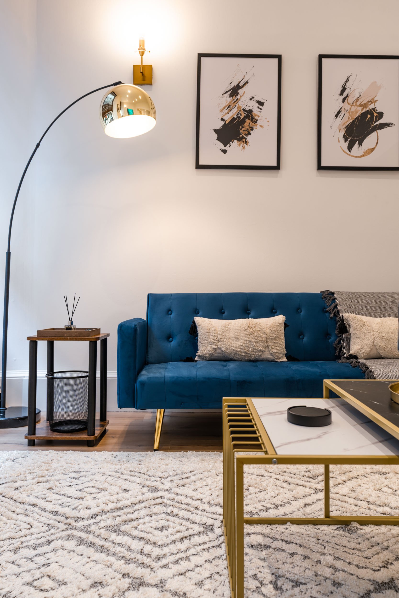 Living room with white walls and modern decor. Features a blue velvet sofa with pillows, a white textured rug, a gold and marble coffee table, a small side table with a reed diffuser, and framed abstract art on the wall. A tall floor lamp with a curv