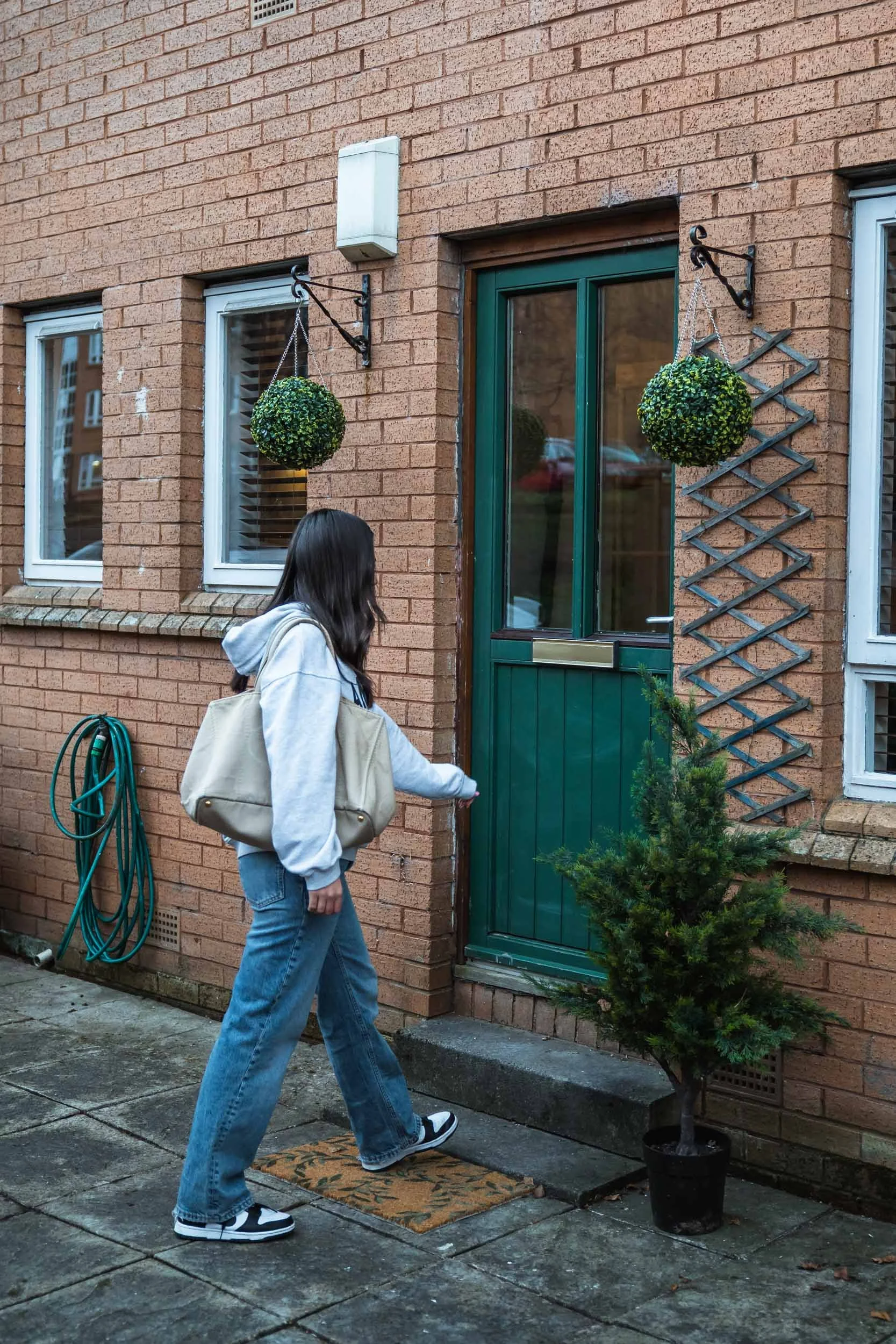 A person with long dark hair, wearing a gray hoodie, baggy jeans, and black and white sneakers, is stepping onto a doorstep and opening a green door with a metal mail slot, outside a brick house. There are hanging plants on either side of the door, a