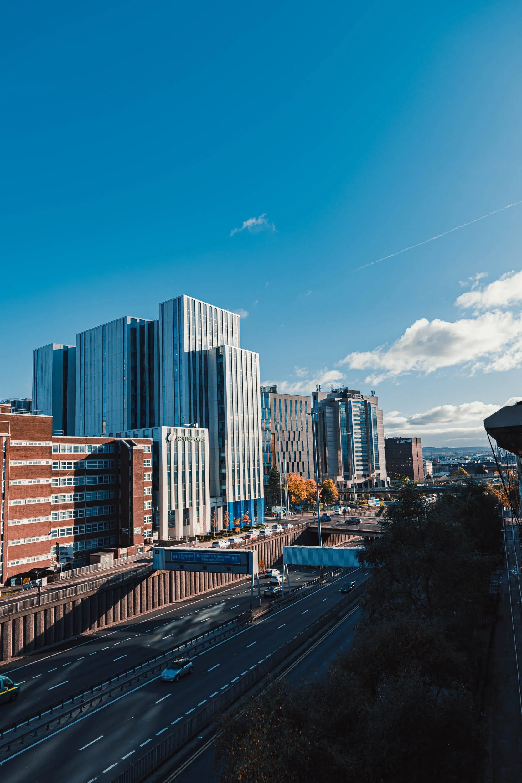 Cityscape with modern skyscrapers, an elevated highway with moving cars, blue sky with scattered clouds, and a few trees in the foreground.