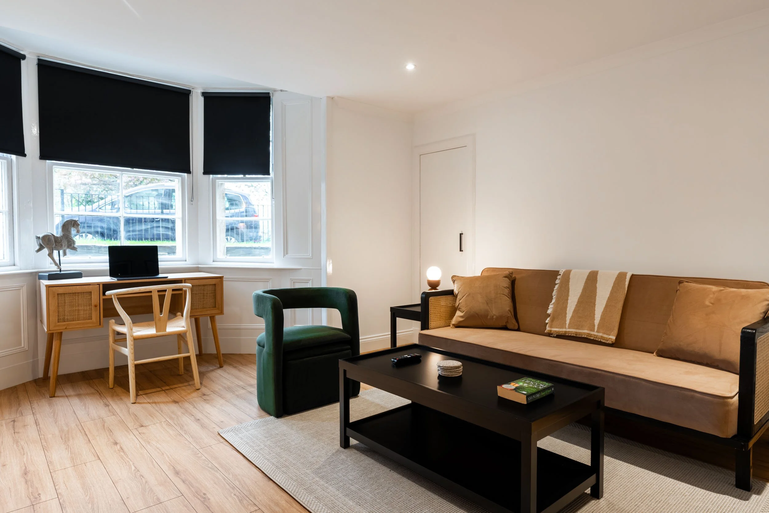 Living room with a beige sofa, green armchair, black coffee table, and a small side table with a lamp. There is a black TV on a wooden console near three windows with black blinds, and a decorative horse figurine on the side.