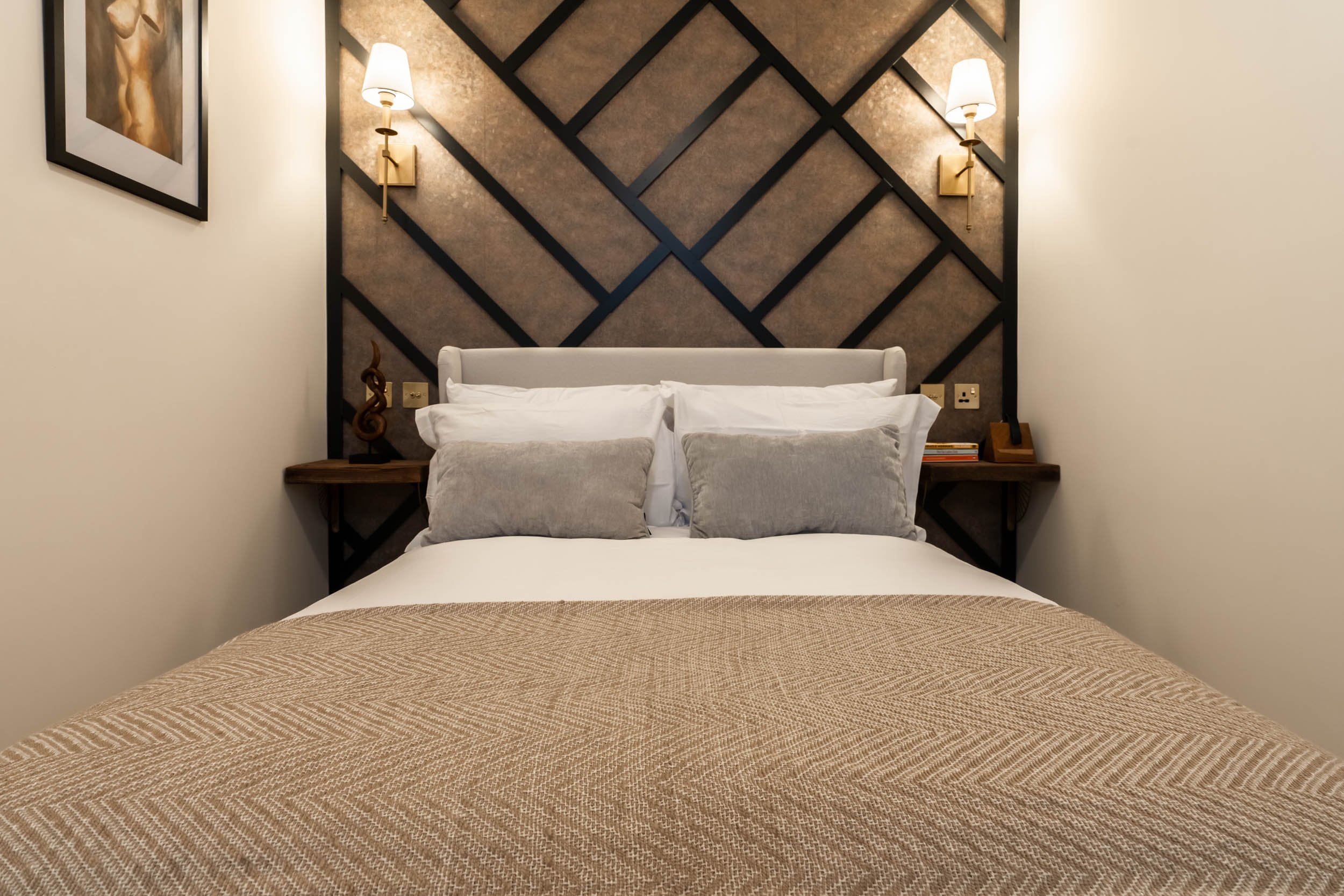 A neatly made bed with white linens, three pillows, and a beige patterned bedspread in a modern bedroom, featuring a decorative geometric wood panel wall behind the bed with two wall-mounted lamps on either side.