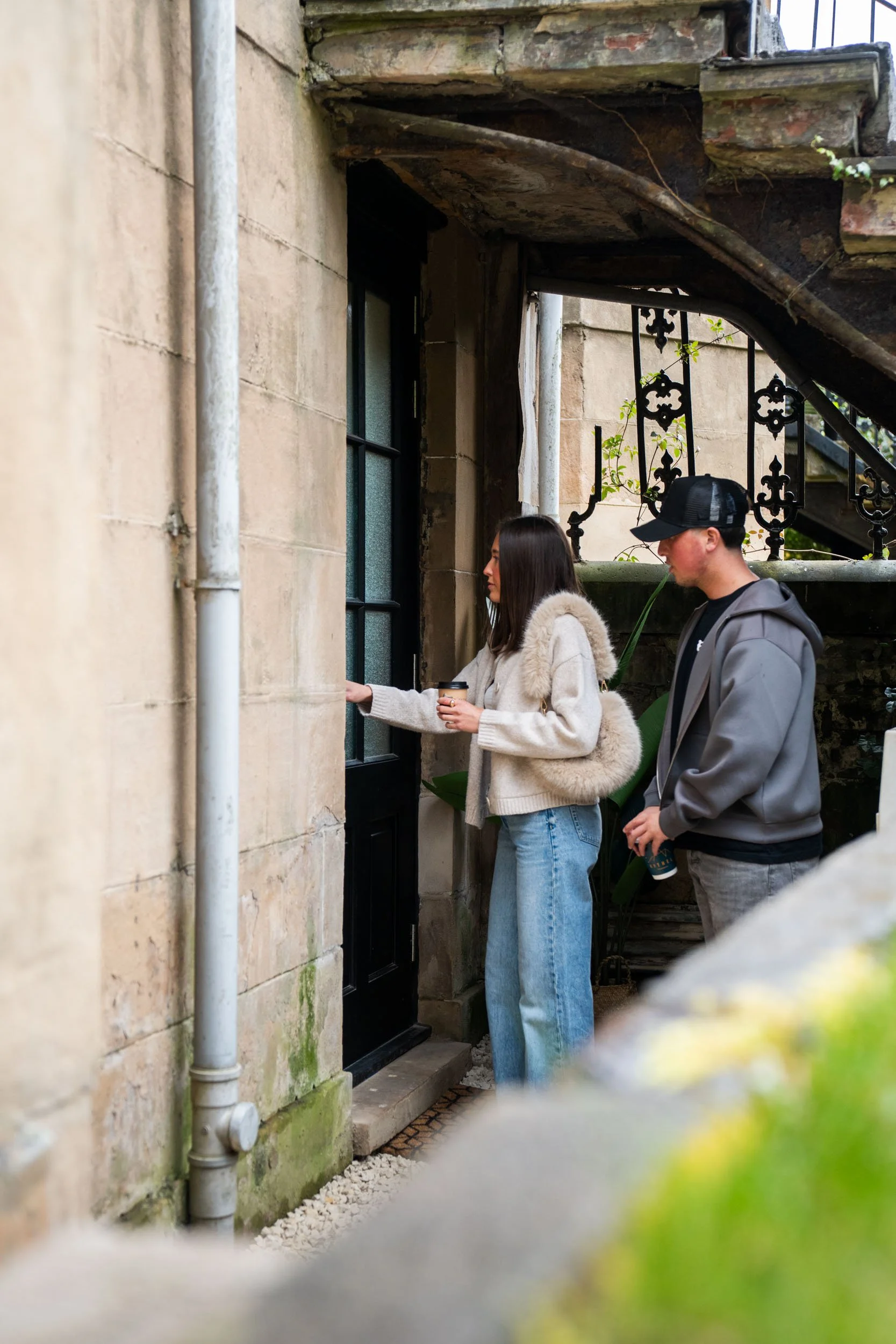 A woman is knocking on a black door while a man stands behind her, holding a beverage, outside a stone building with a staircase and decorative iron railing.