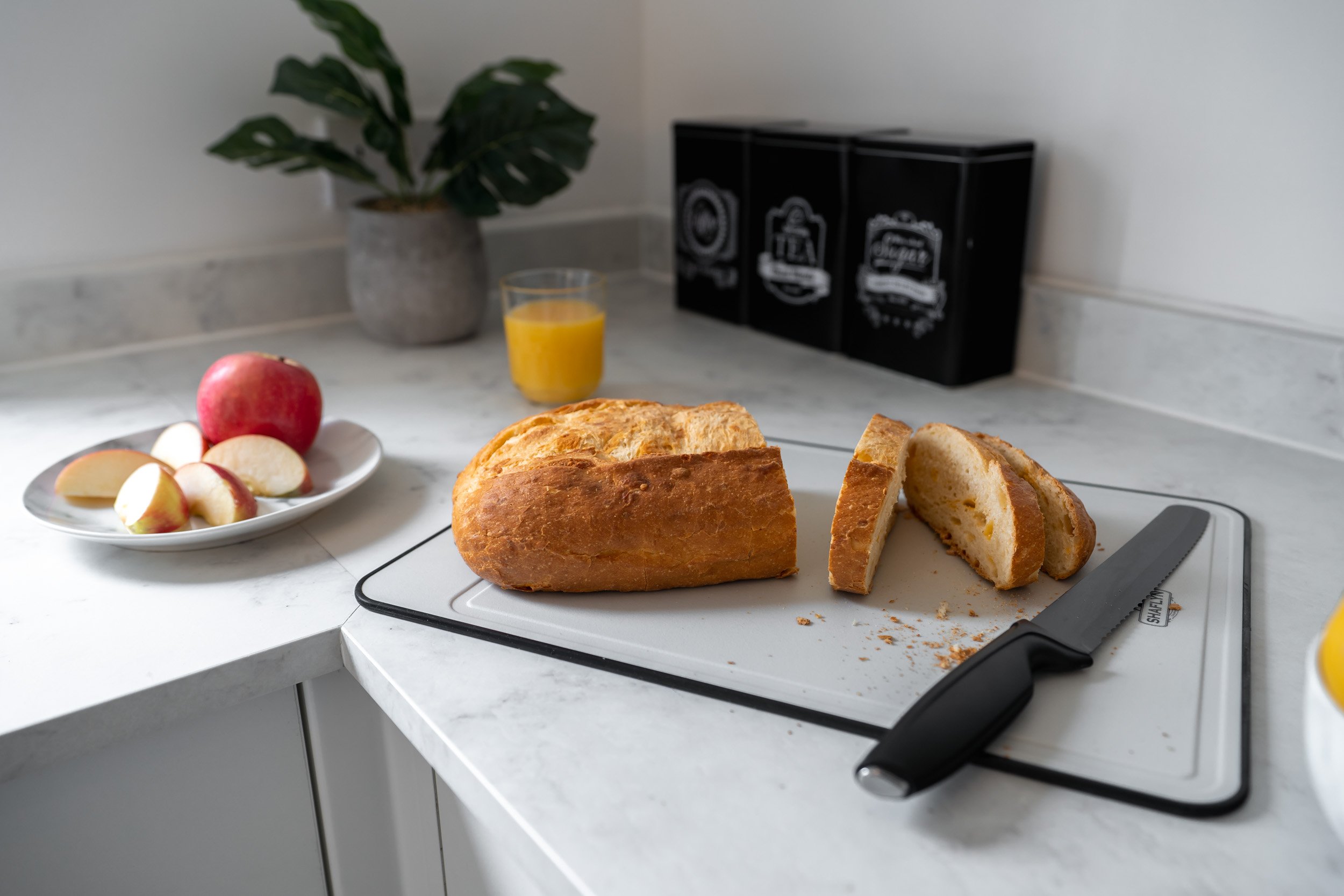Sliced loaf of bread on a cutting board, with a knife. A white plate with a whole apple and sliced apple pieces, a glass of orange juice, and a potted plant in the background.