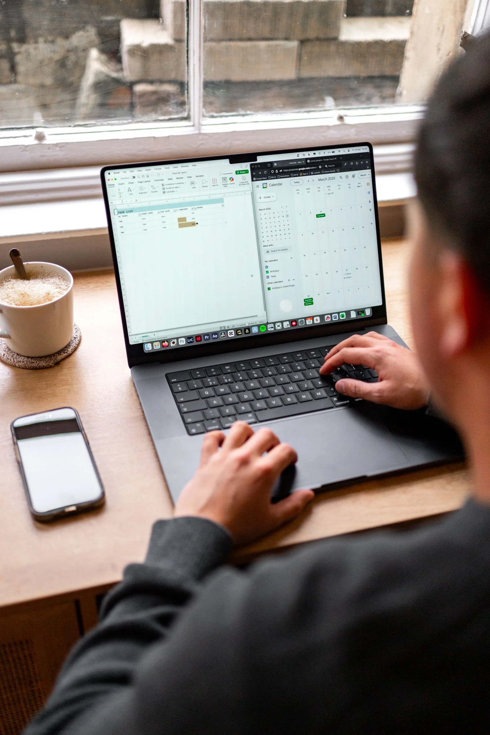 Person working on a laptop with a calendar and spreadsheet open, sitting by a window with a mug of coffee and a smartphone on the desk.