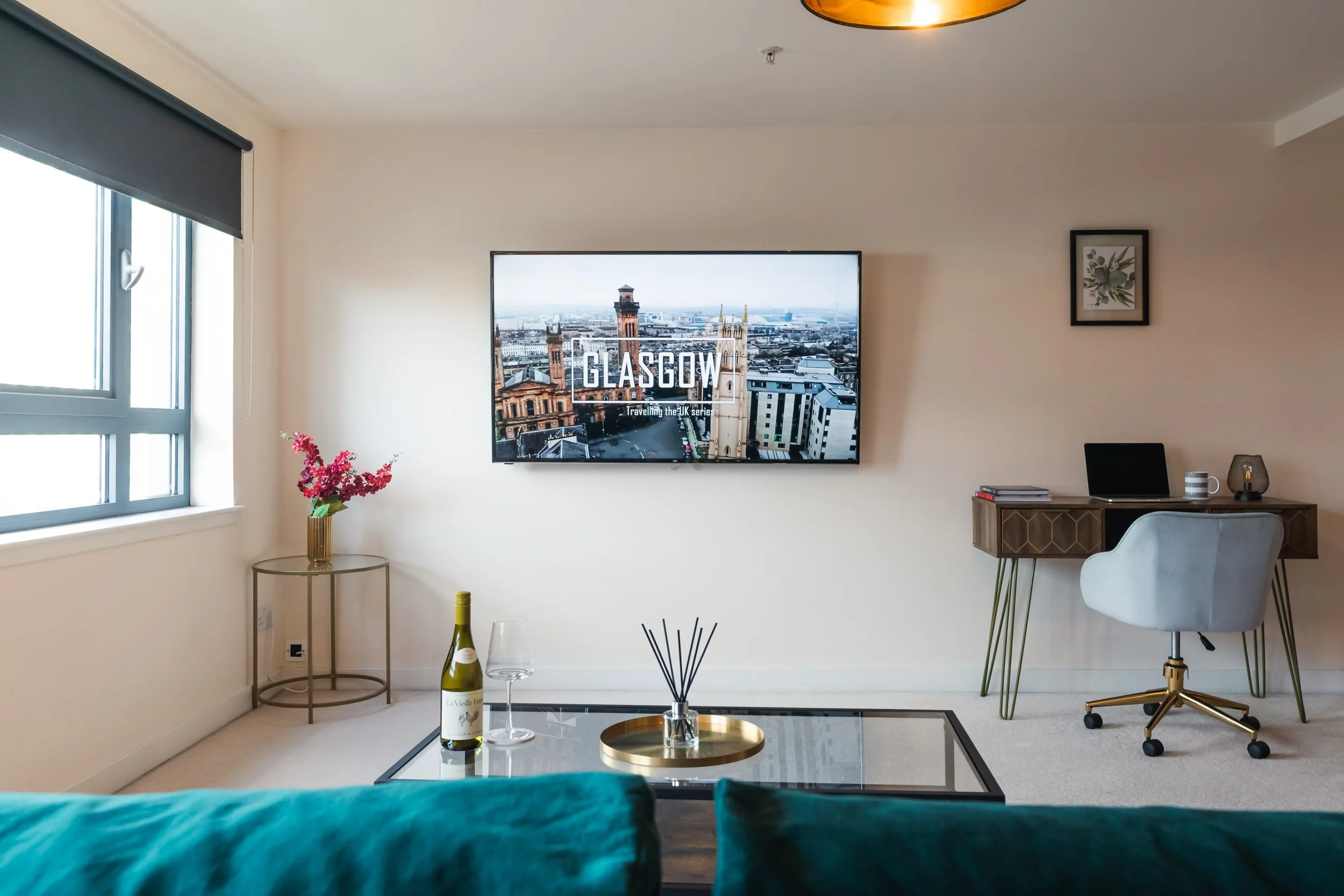 Living room with a wall-mounted TV displaying 'Glasgow' and cityscape, a window with black blinds, a side table with pink flowers, a glass of wine and a wine bottle on a coffee table, a desk with a laptop, books, mug, and decorative items, and a gray