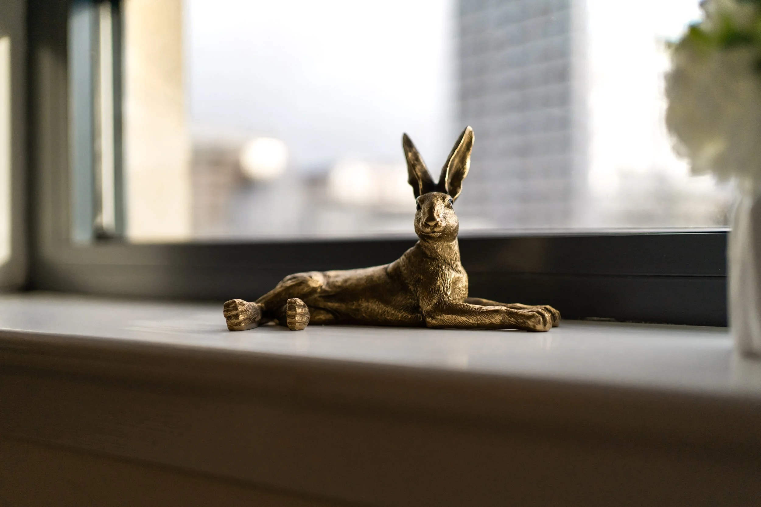 A bronze rabbit figurine lying on a windowsill with a blurred cityscape in the background.
