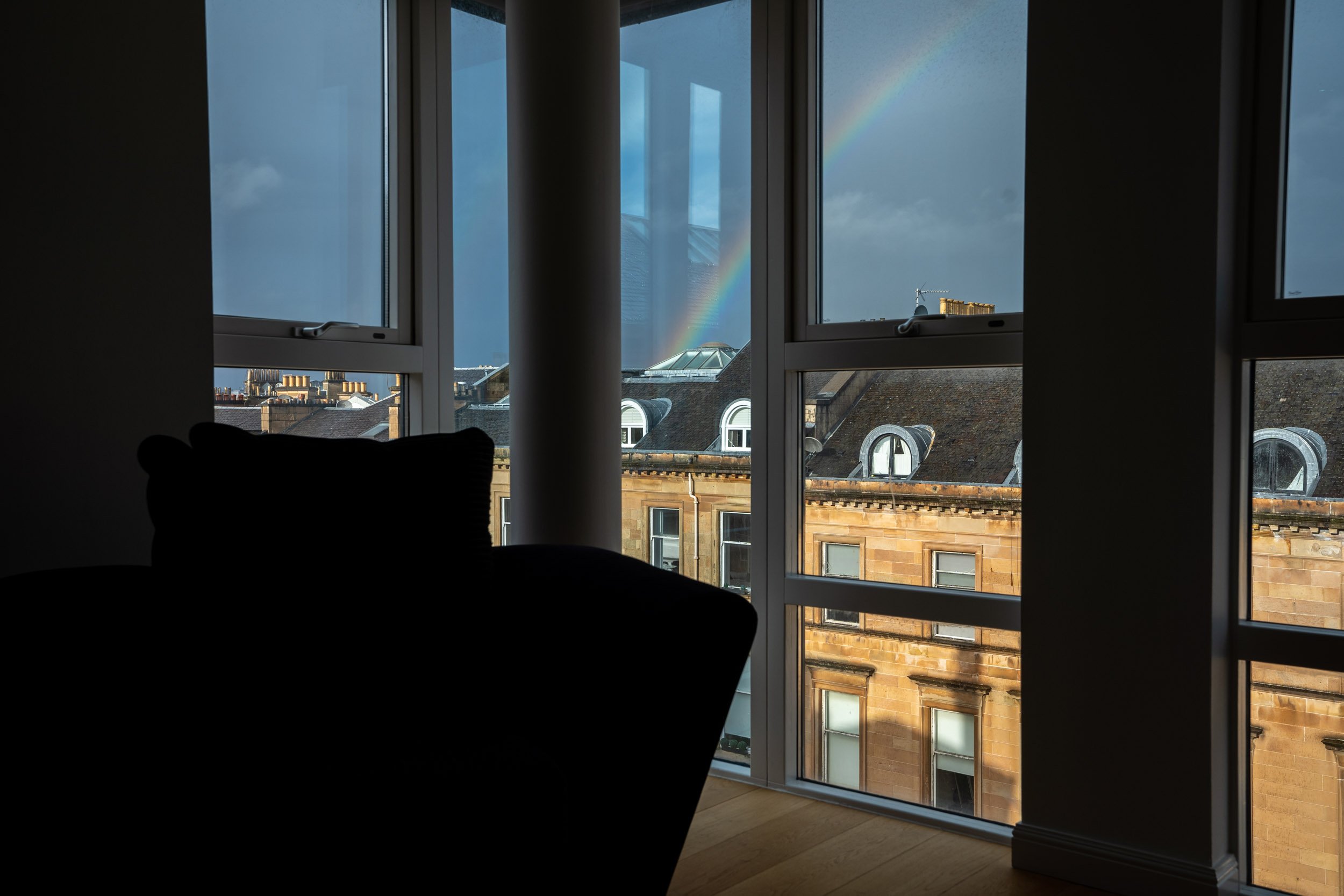 A view from inside a dimly lit room shows a large window with a rainbow visible in the cloudy sky outside. The window frames a cityscape with historic buildings and a construction crane.