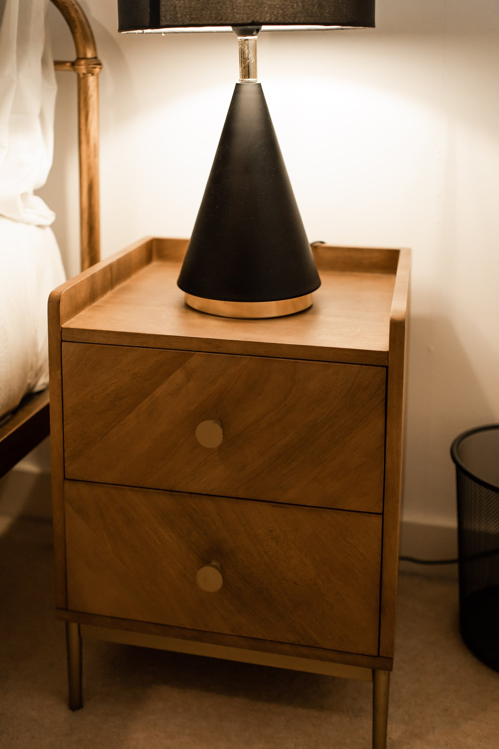 A wooden bedside table with two drawers, each with a round, beige knob, and a black conical table lamp on top, located beside a bed with white bedding.