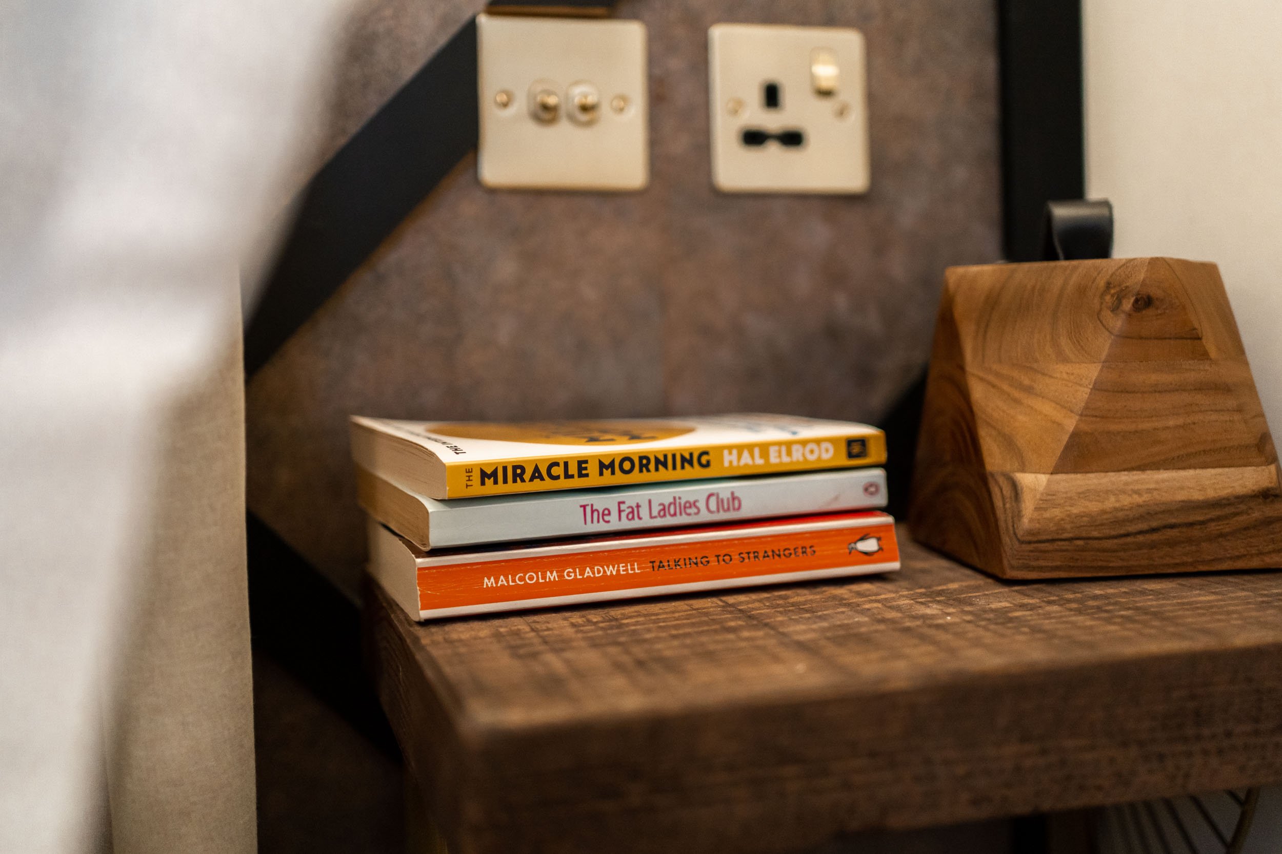 A wooden bedside table with three stacked books, a black lamp, and electrical outlets on the wall behind.