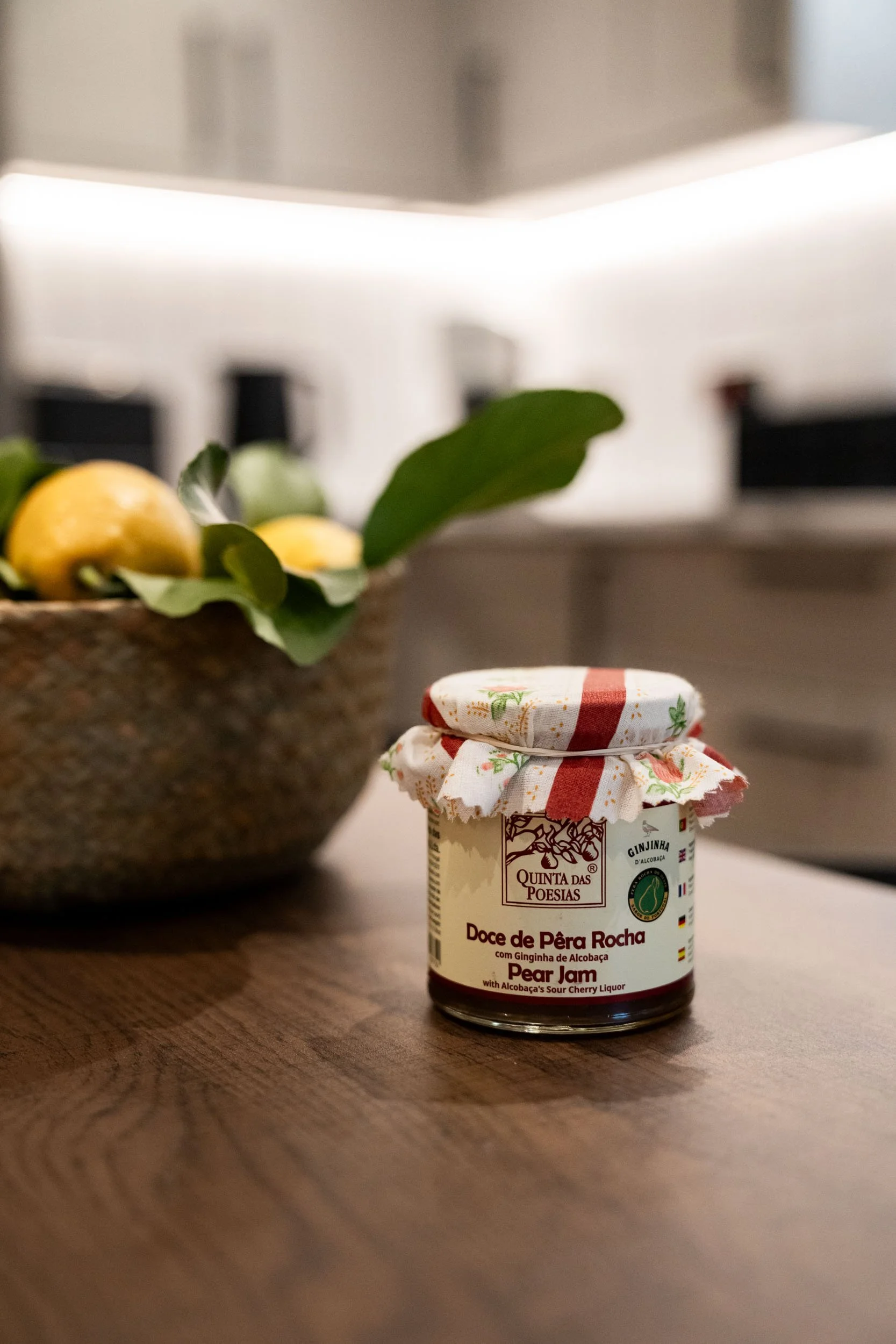 A jar of Pear Jam with alcobaça sour cherry liquor on a wooden table, beside a bowl of apples and green leaves.