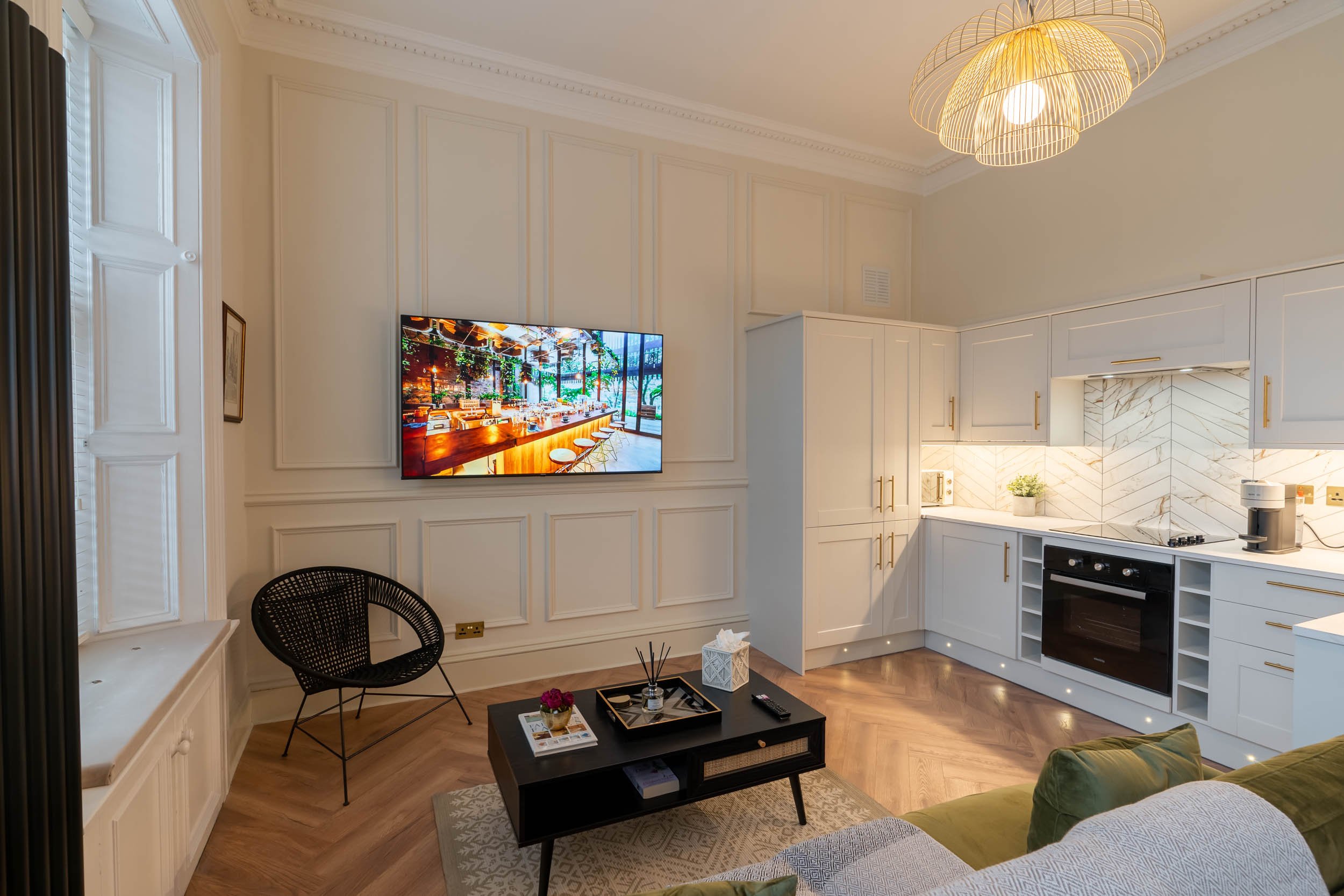 Modern living room and kitchen with white cabinets, black oven, and marble backsplash. A black chair, coffee table with decor, and a wall-mounted TV are visible, with wooden flooring and a large gold and white pendant light.