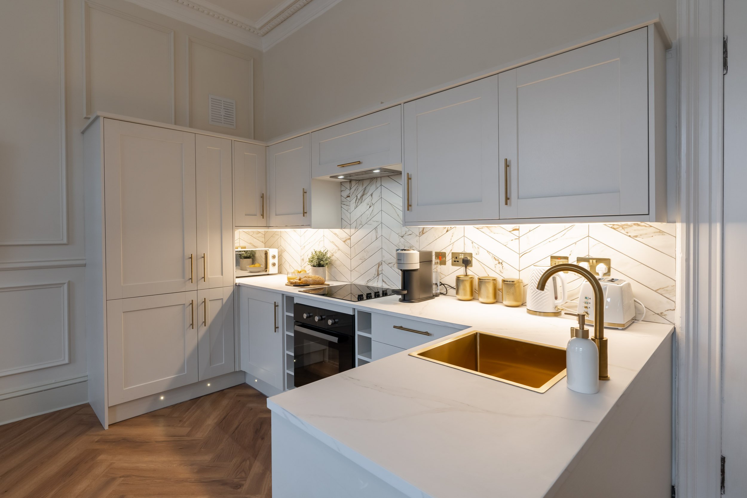 Modern kitchen with white cabinets, gold handles, marble backsplash, gold sink, and wooden flooring, with small appliances and decor on the counter.
