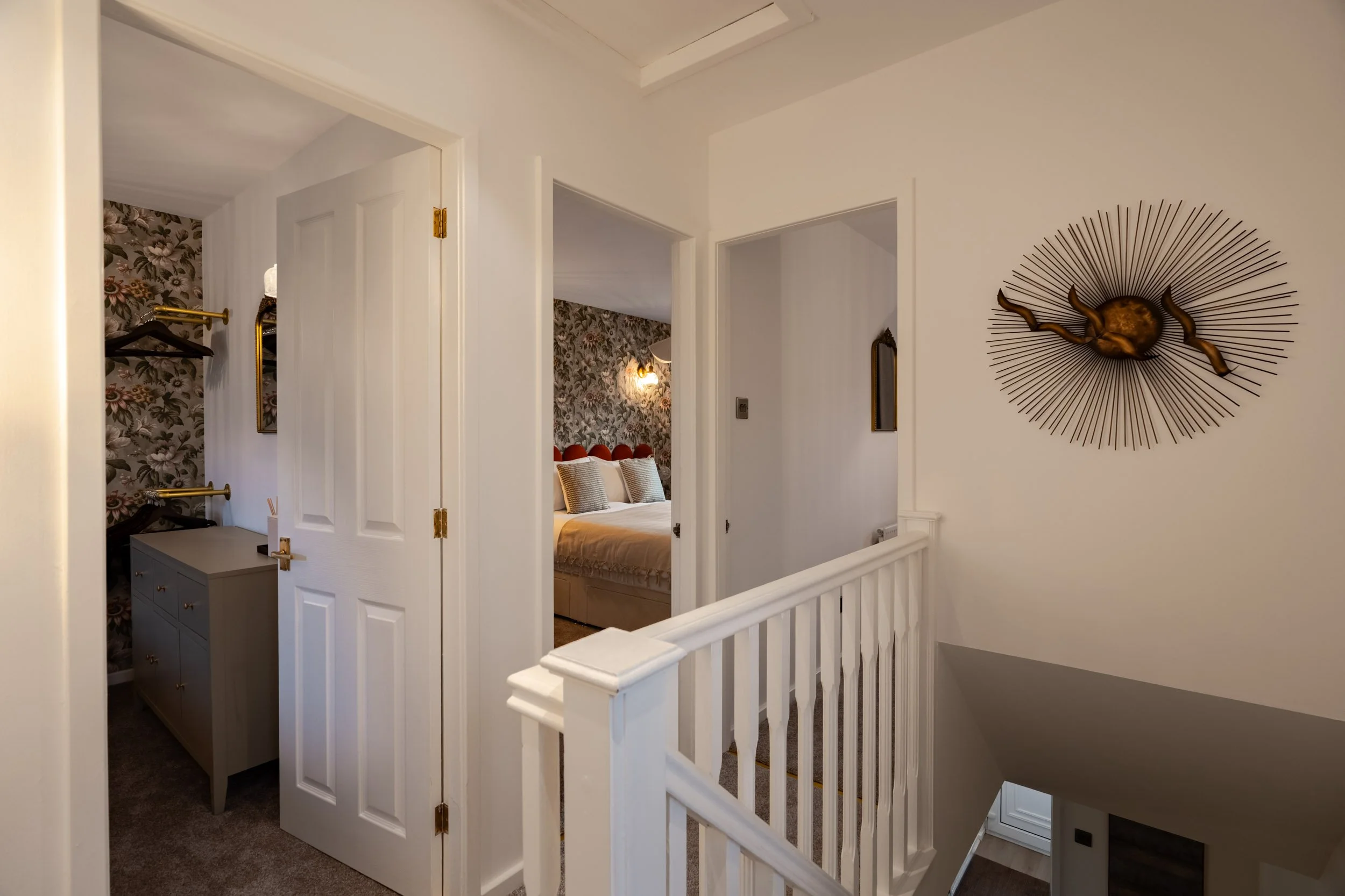 View of an upstairs hallway with open doors leading to bedrooms, featuring white walls, a stair railing, a decorative wall art piece, and wallpapered bedrooms with floral patterns.