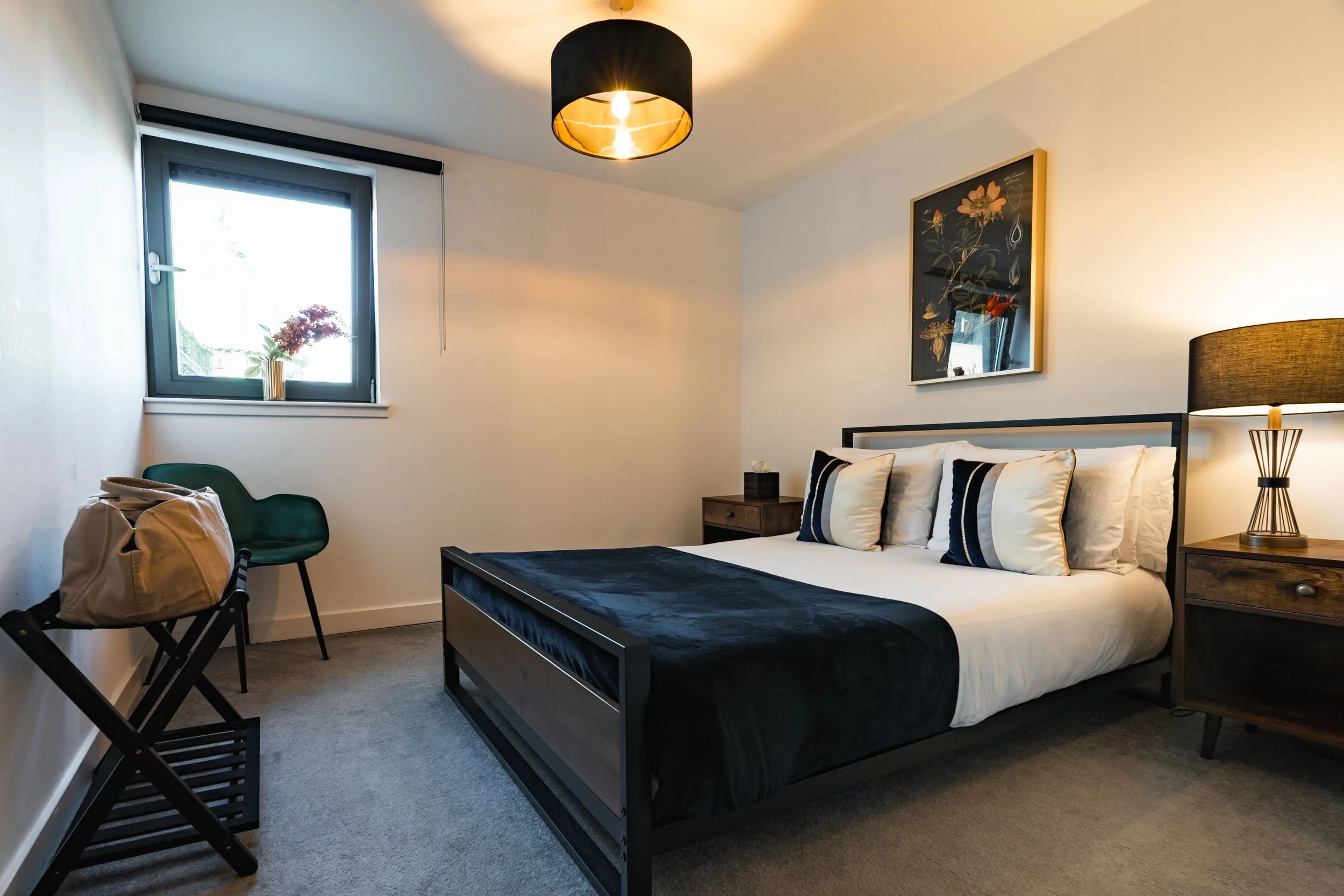 Modern bedroom with a black metal bed frame, white bedding, three decorative pillows, a nightstand with a lamp, a small window with a plant on the sill, and a black ceiling light