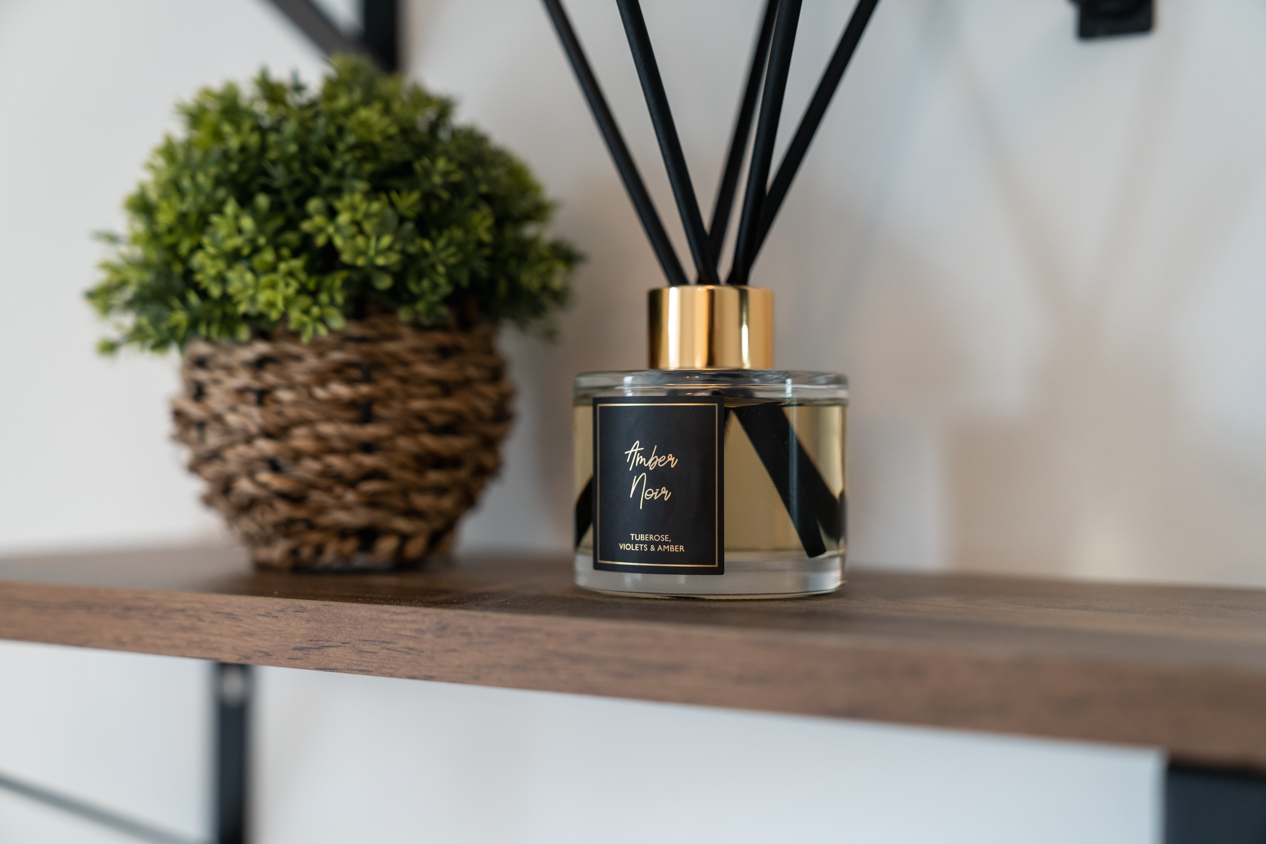 A decorative reed diffuser with black reeds and a black and gold bottle label reading "Amber Noir," placed on a wooden surface next to a potted green plant in a woven basket.