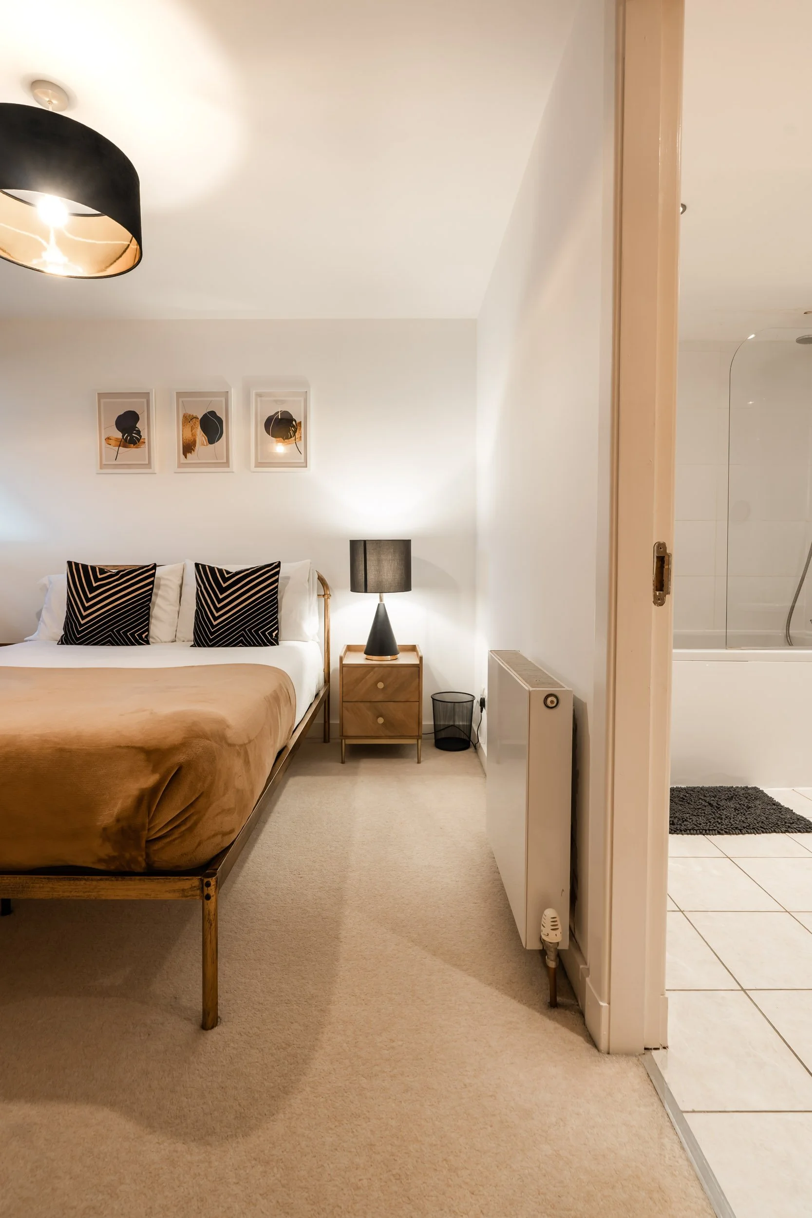 A bedroom with a bed, black and white pillows, a wooden nightstand, a black table lamp, and framed artwork on the white wall. A doorway leads to a bathroom with a bathtub, shower, gray rug, and white tiled floor.