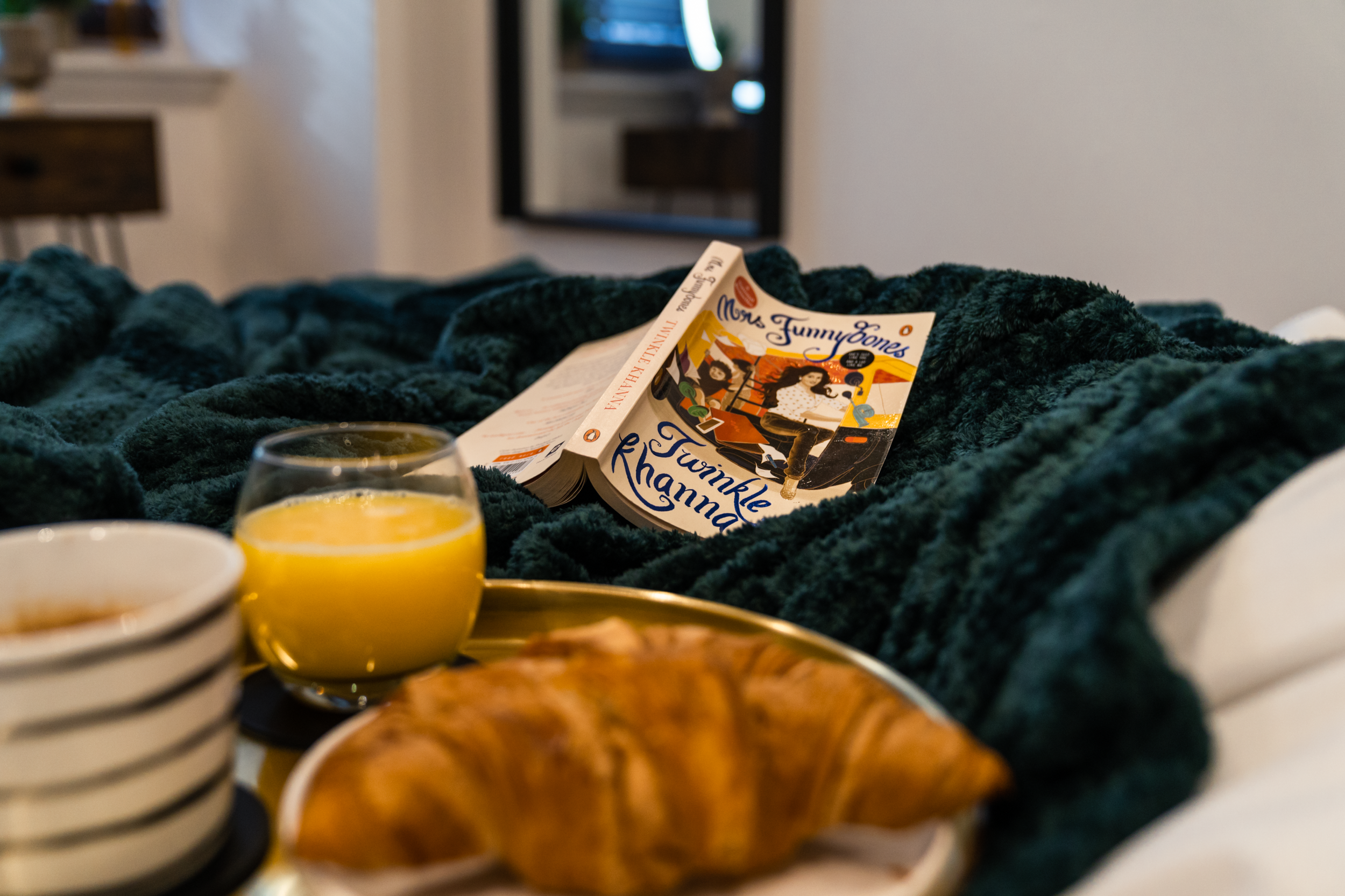 A cozy bed with a dark green blanket, a book titled Twinkle Rhanna by Moms Funny Bones, a glass of orange juice, a croissant on a plate, and two cups of coffee in striped cups.