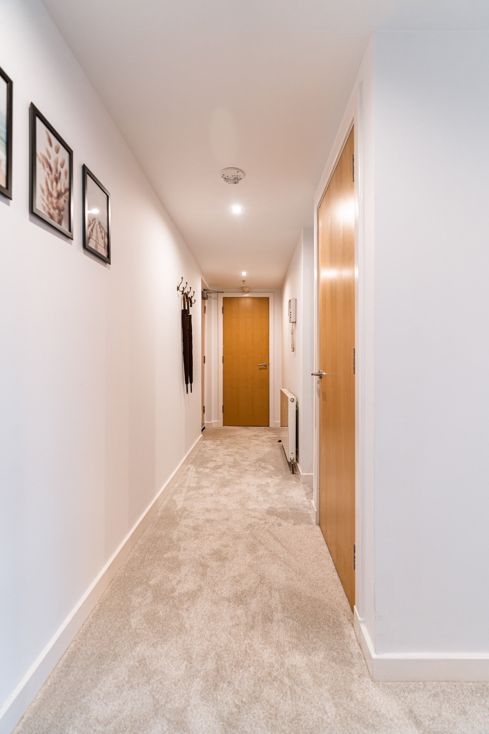 A hallway with white walls, beige carpet, and wooden doors. Framed pictures hang on the left wall, and a coat rack is mounted on the left wall. Recessed ceiling lights illuminate the corridor, which ends with a closed door.