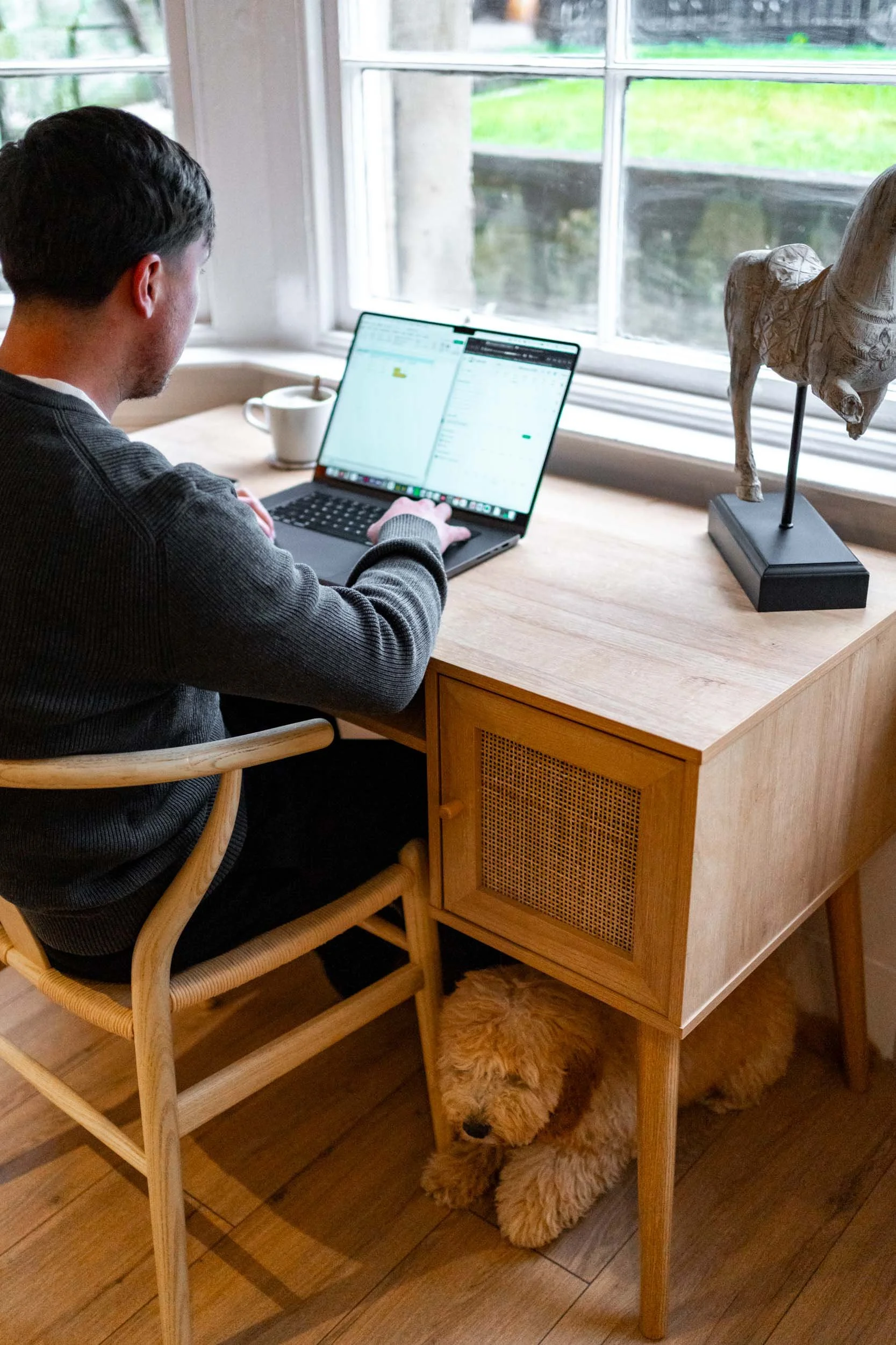 A person working on a laptop at a wooden desk near a window, with a ceramic horse sculpture and a mug nearby. A dog is lying underneath the desk.