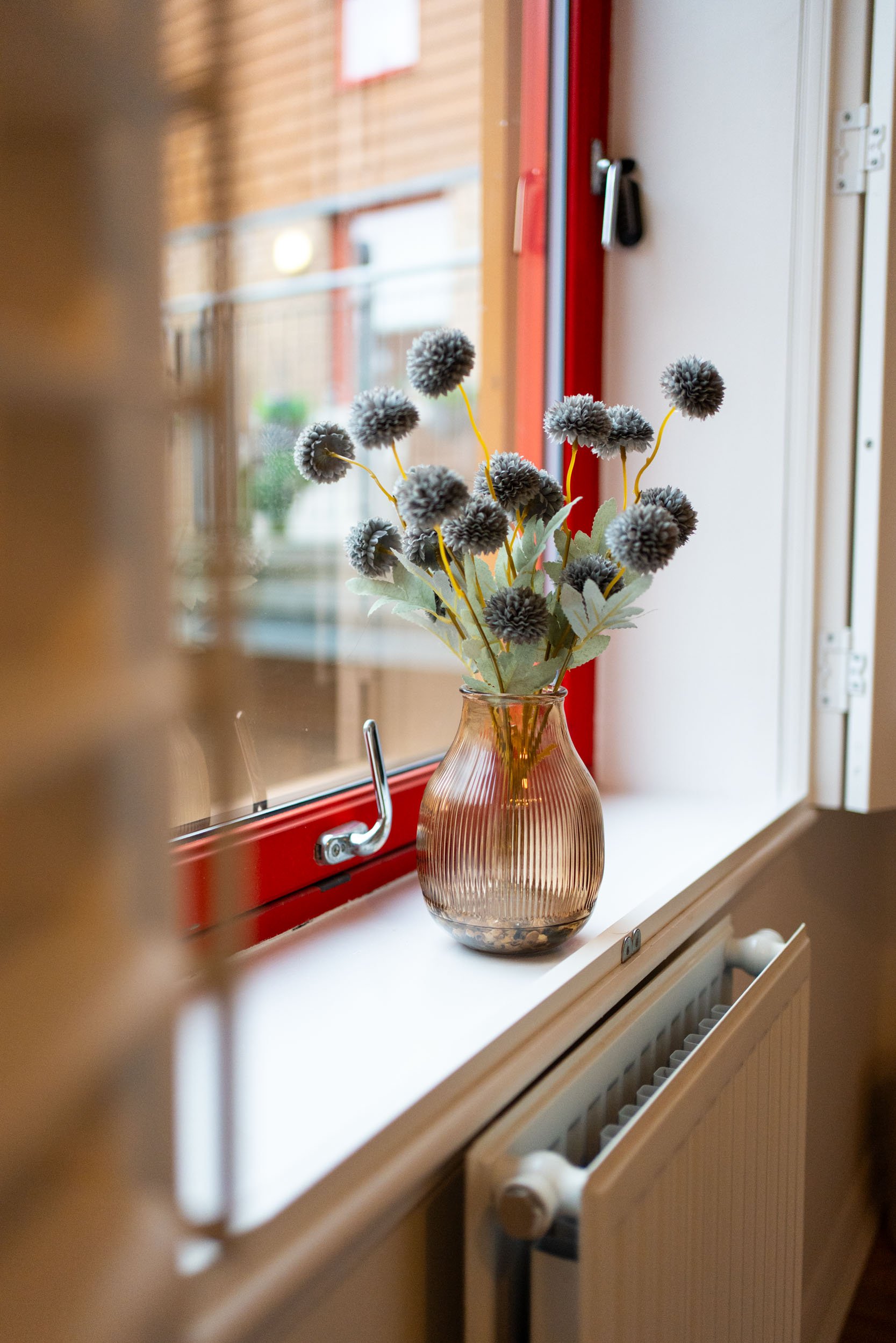 A vase with artificial gray pom-pom flowers and green leaves on a white windowsill inside a room, with a radiator below the window and a red window frame.