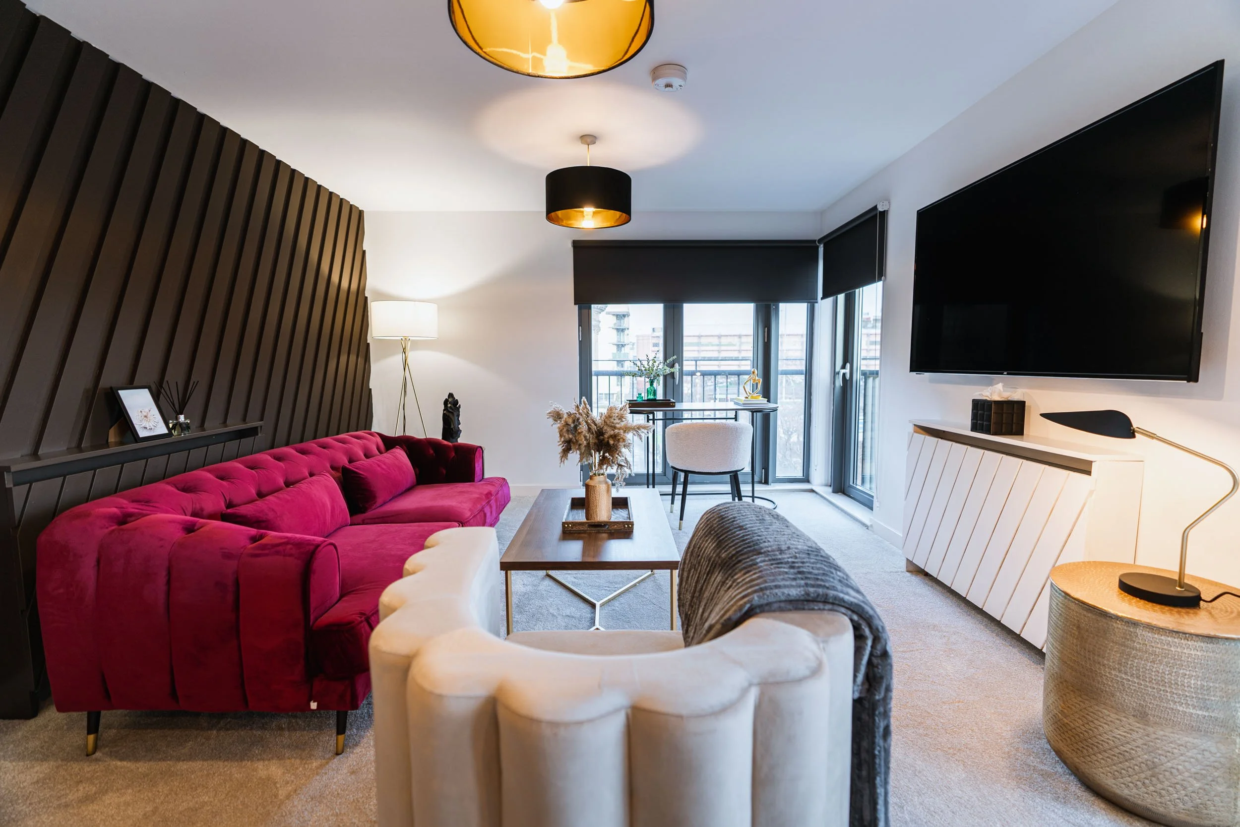 Modern living room with a red velvet sofa, a beige armchair, a wooden coffee table with a vase, a large TV mounted on the wall, and a small desk near the window with a chair. The room has black, white, and neutral decor with natural light coming from