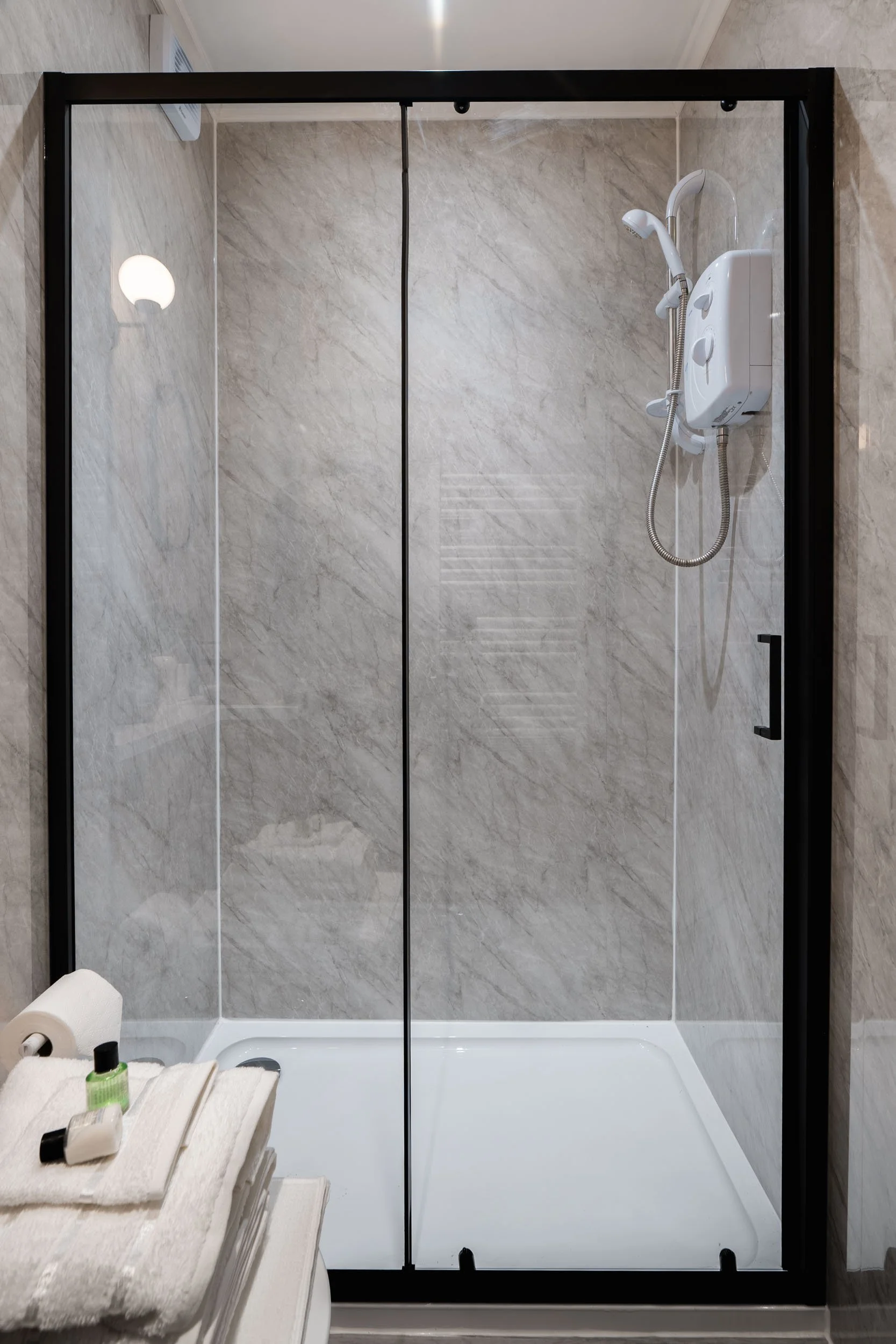 Empty shower with glass doors, mounted shower head with control panel, and a shelf with towels and toiletries outside the shower area.