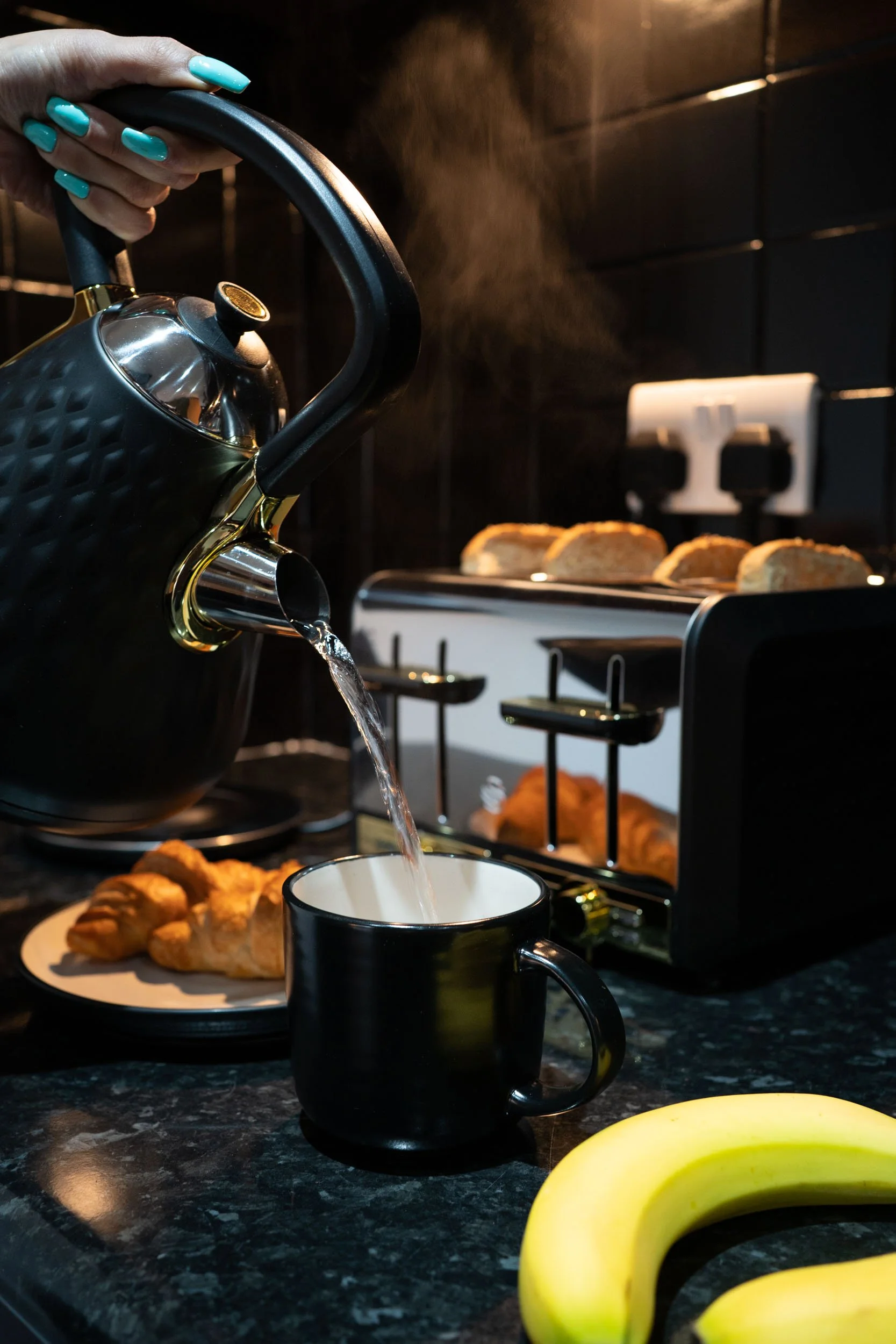A hand with turquoise painted nails pouring hot water from a black kettle into a black mug on a black kitchen countertop, with a toaster, croissants, bananas, and bread in the background.