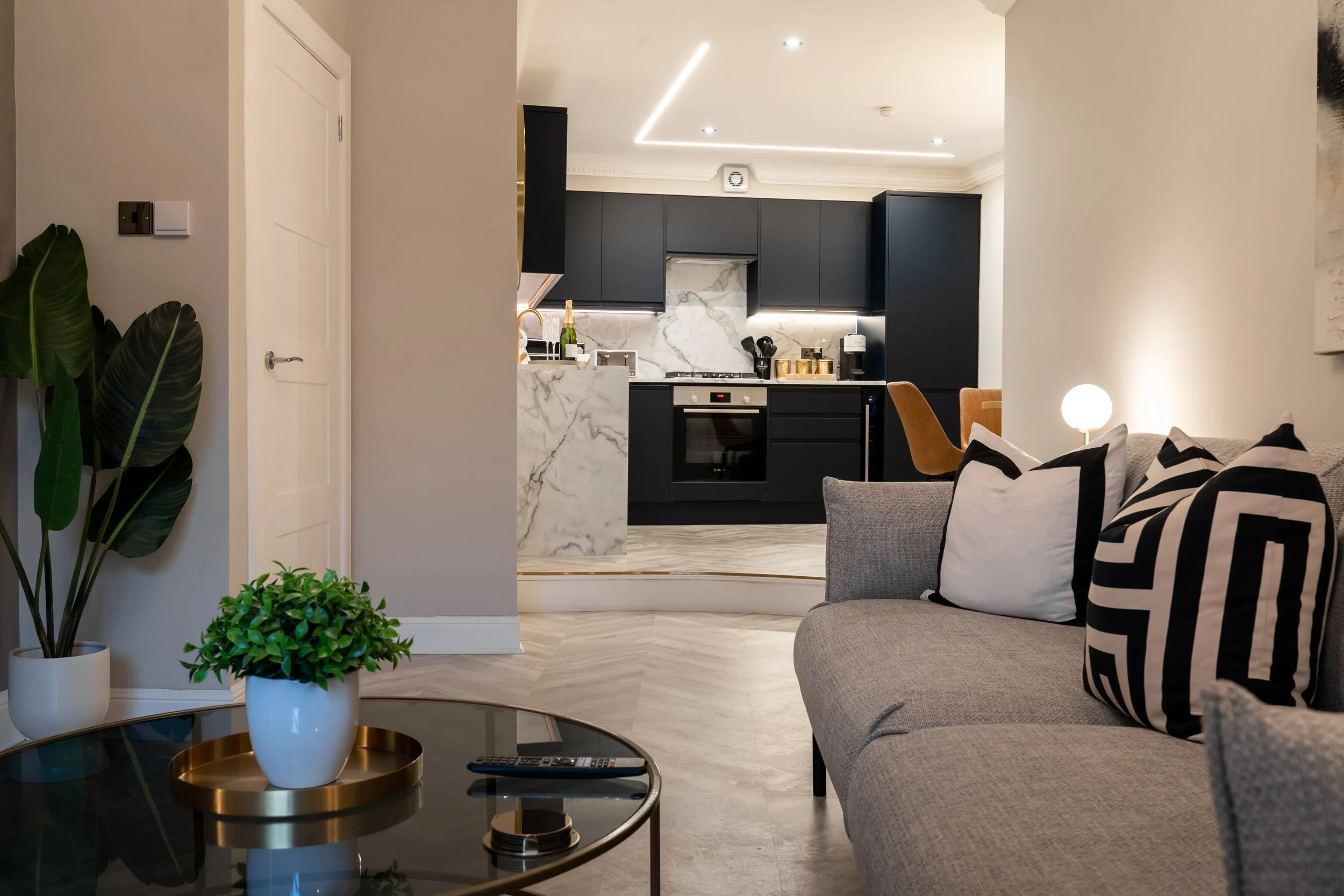 Modern living room with a beige sofa and black and white pillows, a round glass coffee table with a plant and remote control, green plants in white pots, and an open kitchen with black cabinets and marble countertops in the background.