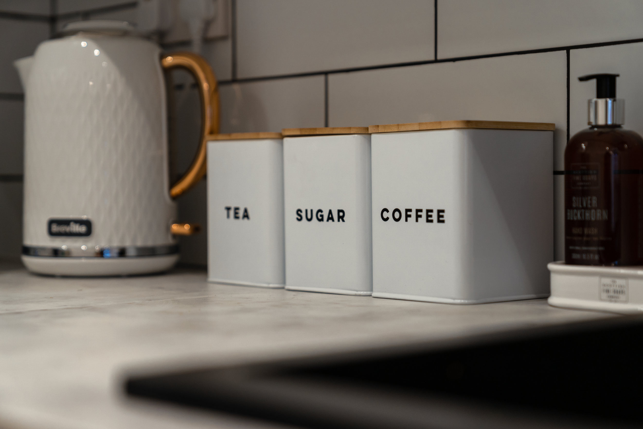Kitchen countertop with white containers labeled 'Tea,' 'Sugar,' and 'Coffee,' an electric kettle on the left, and a brown soap dispenser on the right.