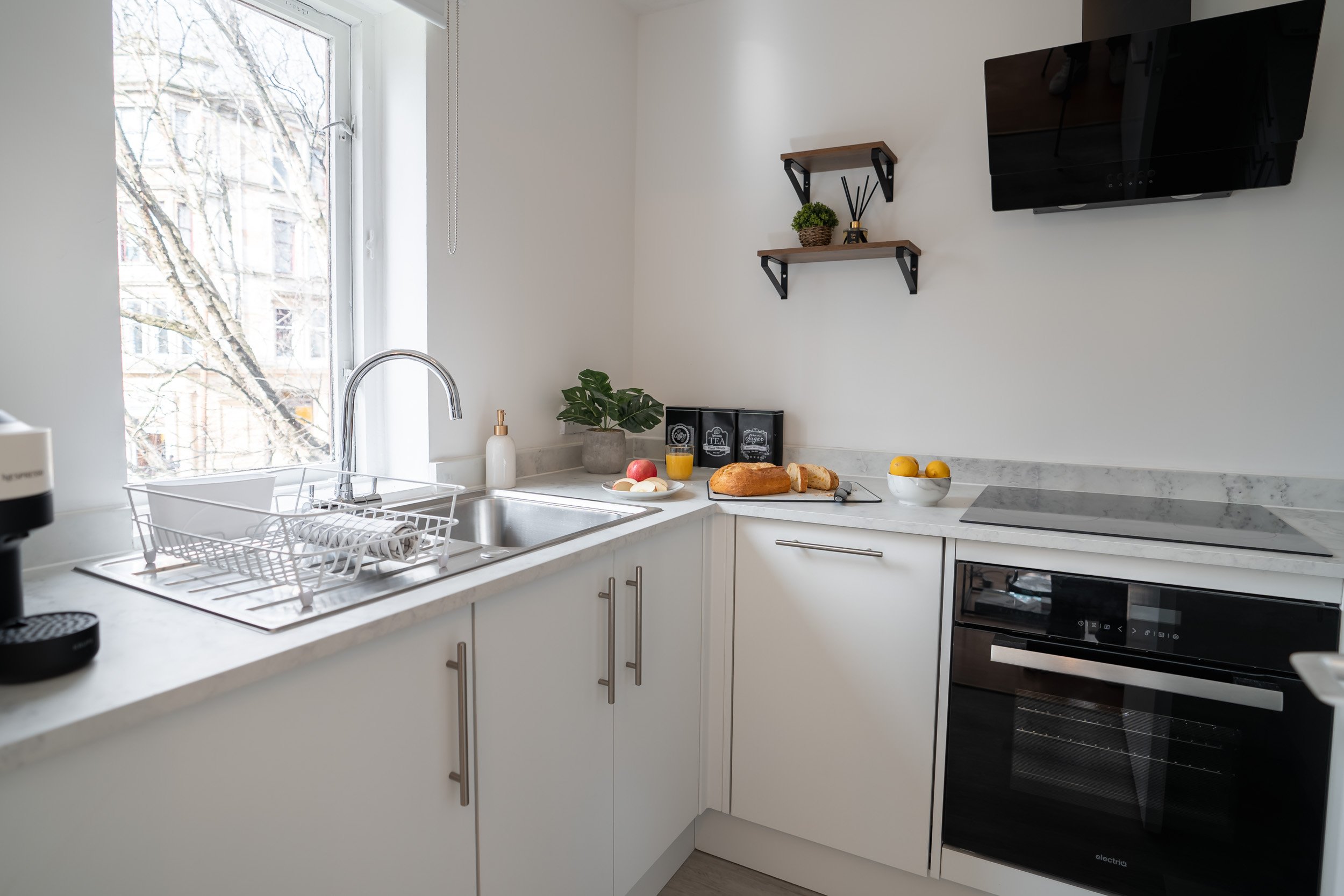 Modern kitchen with white cabinets, marble countertops, a sink under a window, a black oven, and a wall-mounted television. Counter has bread, eggs, and a bowl of lemons.