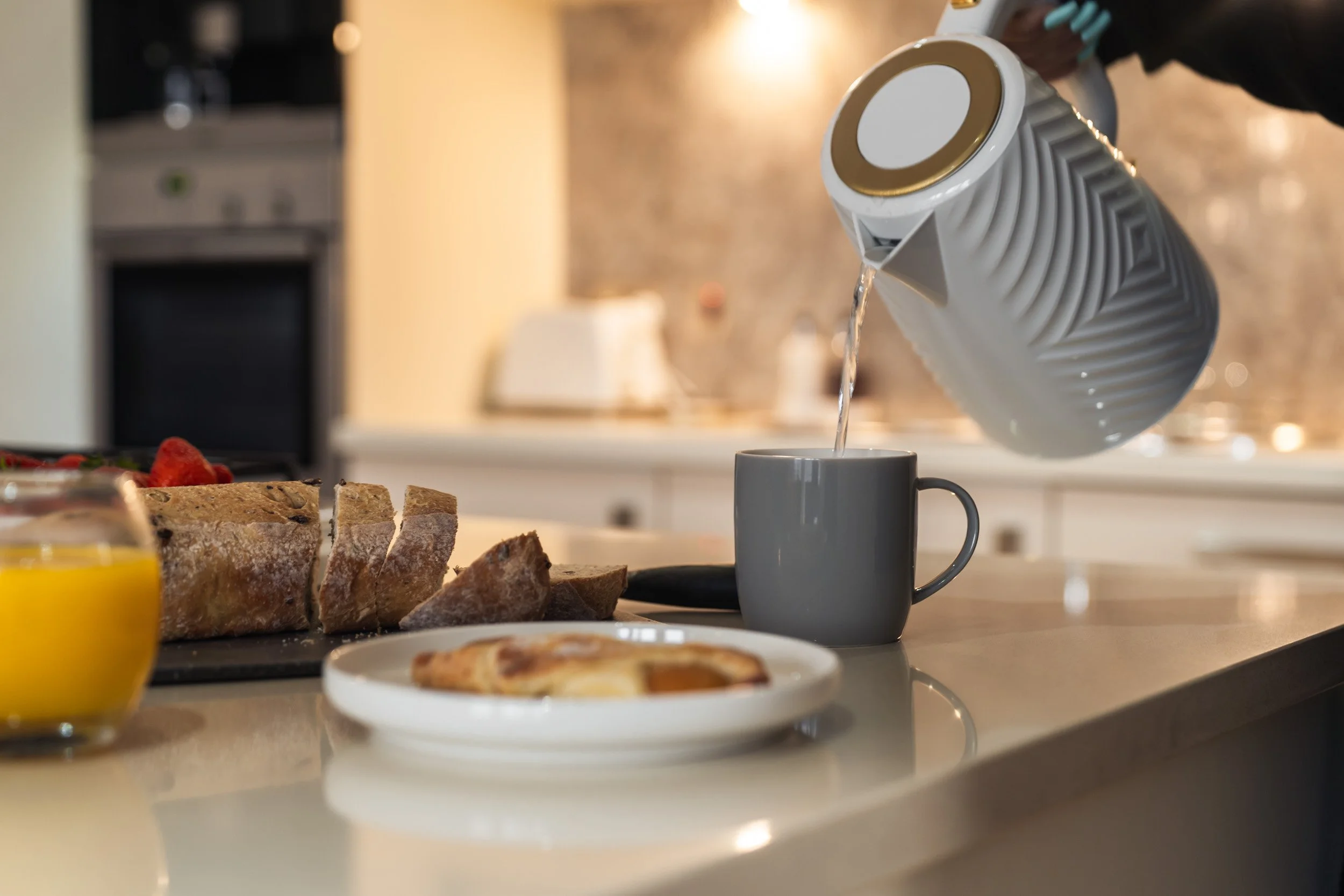 Pouring hot water into a gray mug from a white kettle with gold accents on a kitchen countertop. In the background, there is sliced bread, a plate with a pastry, and a glass of orange juice.