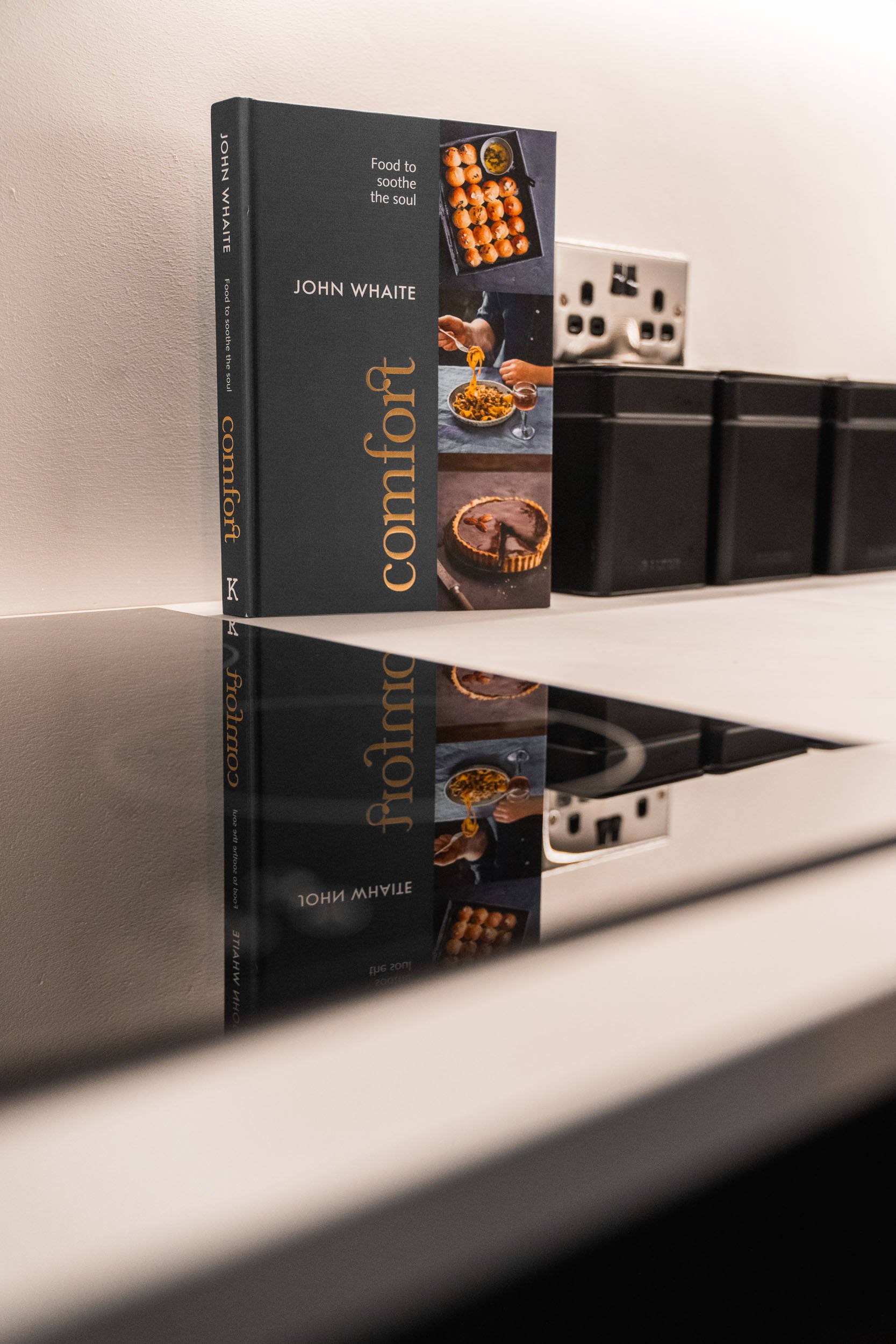A cookbook titled "Comfort" by John Whate standing upright on a kitchen counter, with its reflection visible on the shiny surface below. The cookbook cover features images of a tray of baked goods, a person serving food, and a sliced pie.