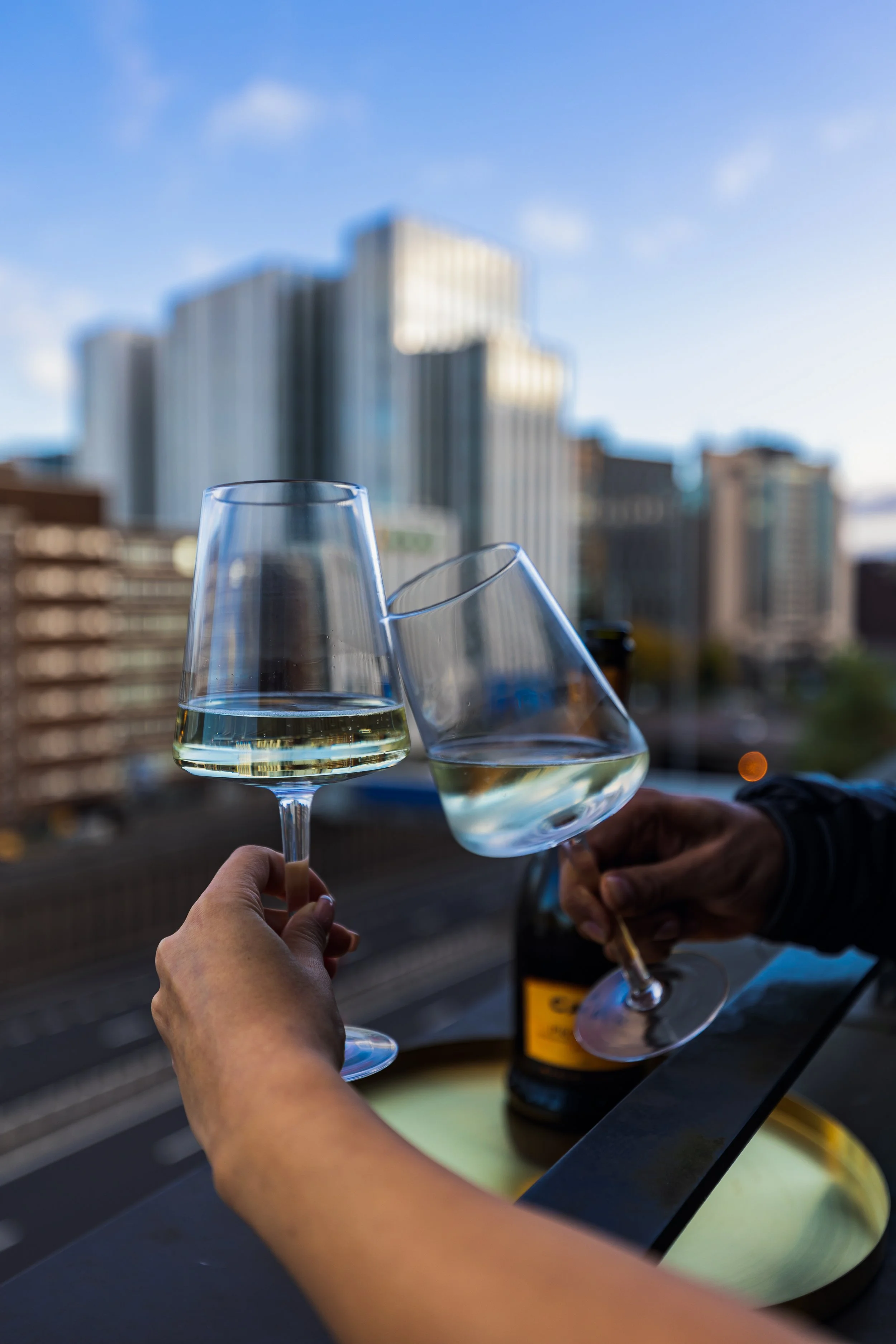 Two people clinking glasses of white wine on a balcony, with city buildings in the background.
