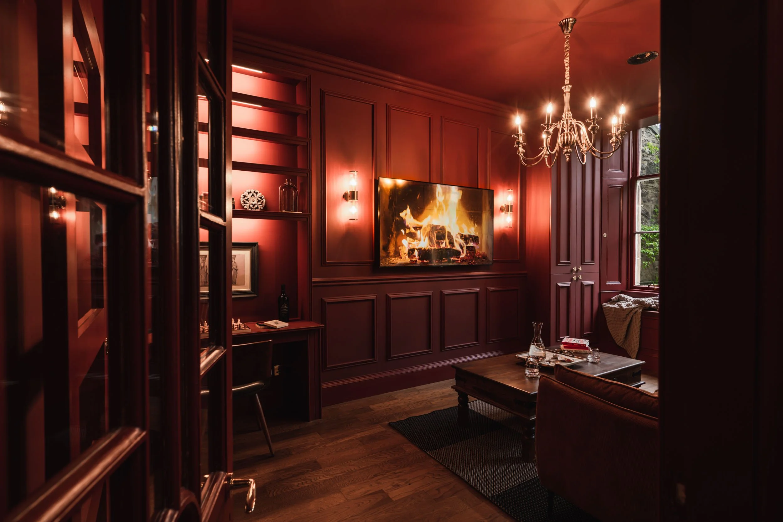 Cozy living room with red walls, a chandelier, wall-mounted TV showing a fireplace, a window with green trees outside, and a wooden coffee table with glassware and books.