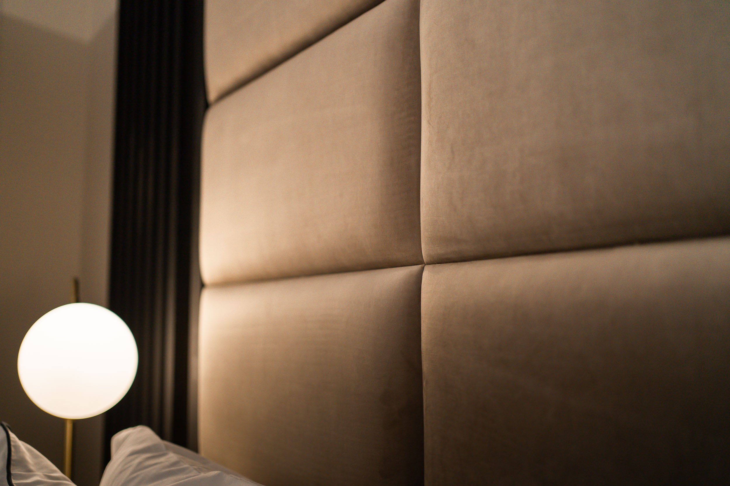 Close-up view of a padded beige headboard in a bedroom, with a white bedside lamp on the left side.