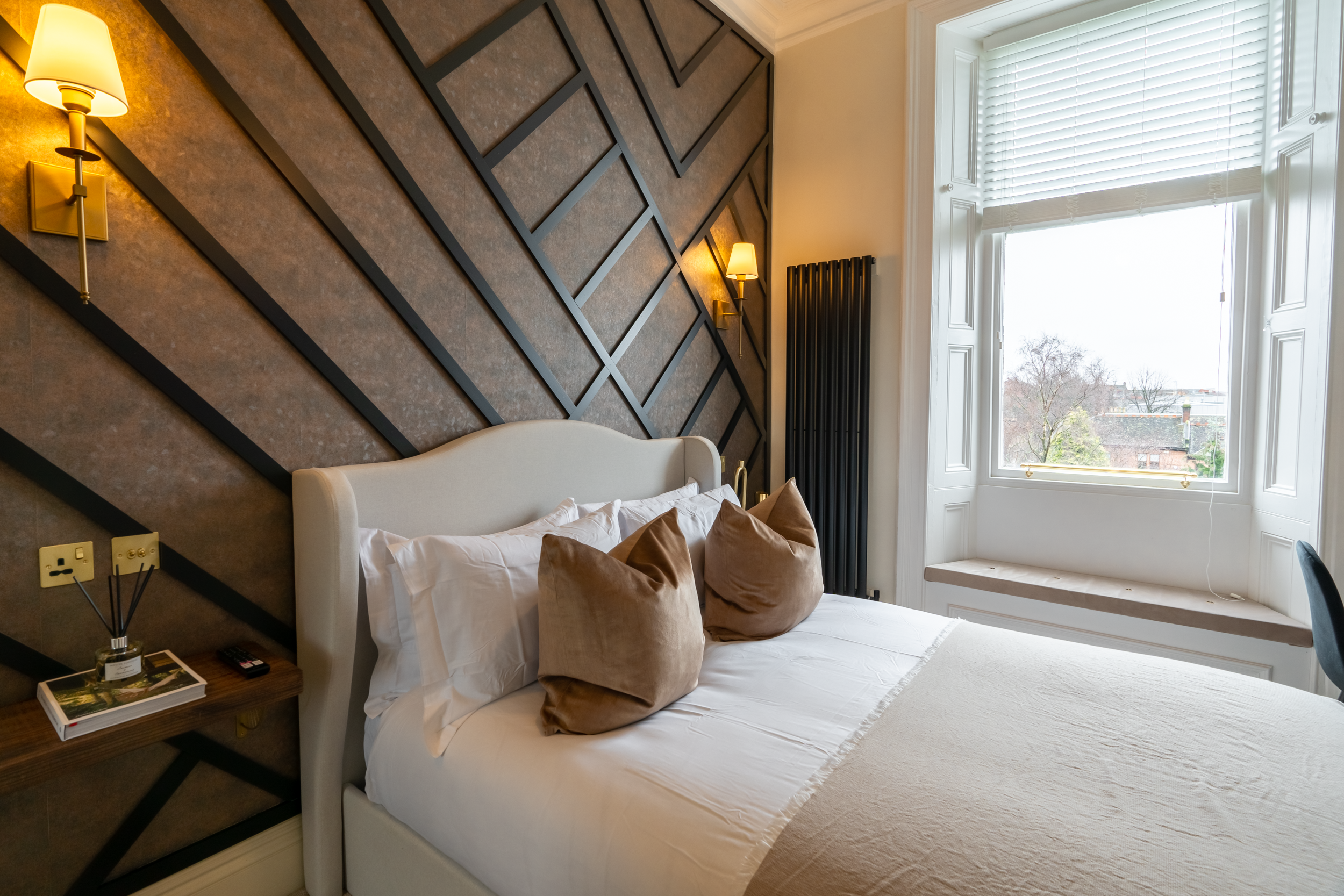A neatly made bed with a white headboard, brown pillows, and a beige blanket, against a geometric patterned accent wall in a bedroom with a large window overlooking trees and buildings.