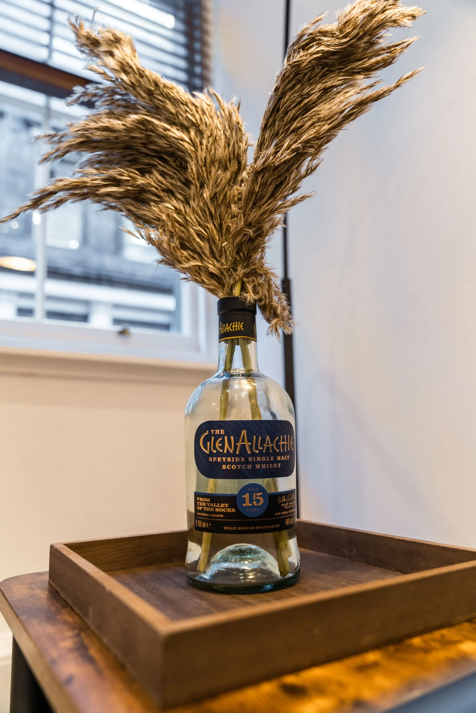 A bottle of Glen Allachie Speyside single malt Scotch whisky with dried pampas grass stems inside, placed on a wooden tray near a window.