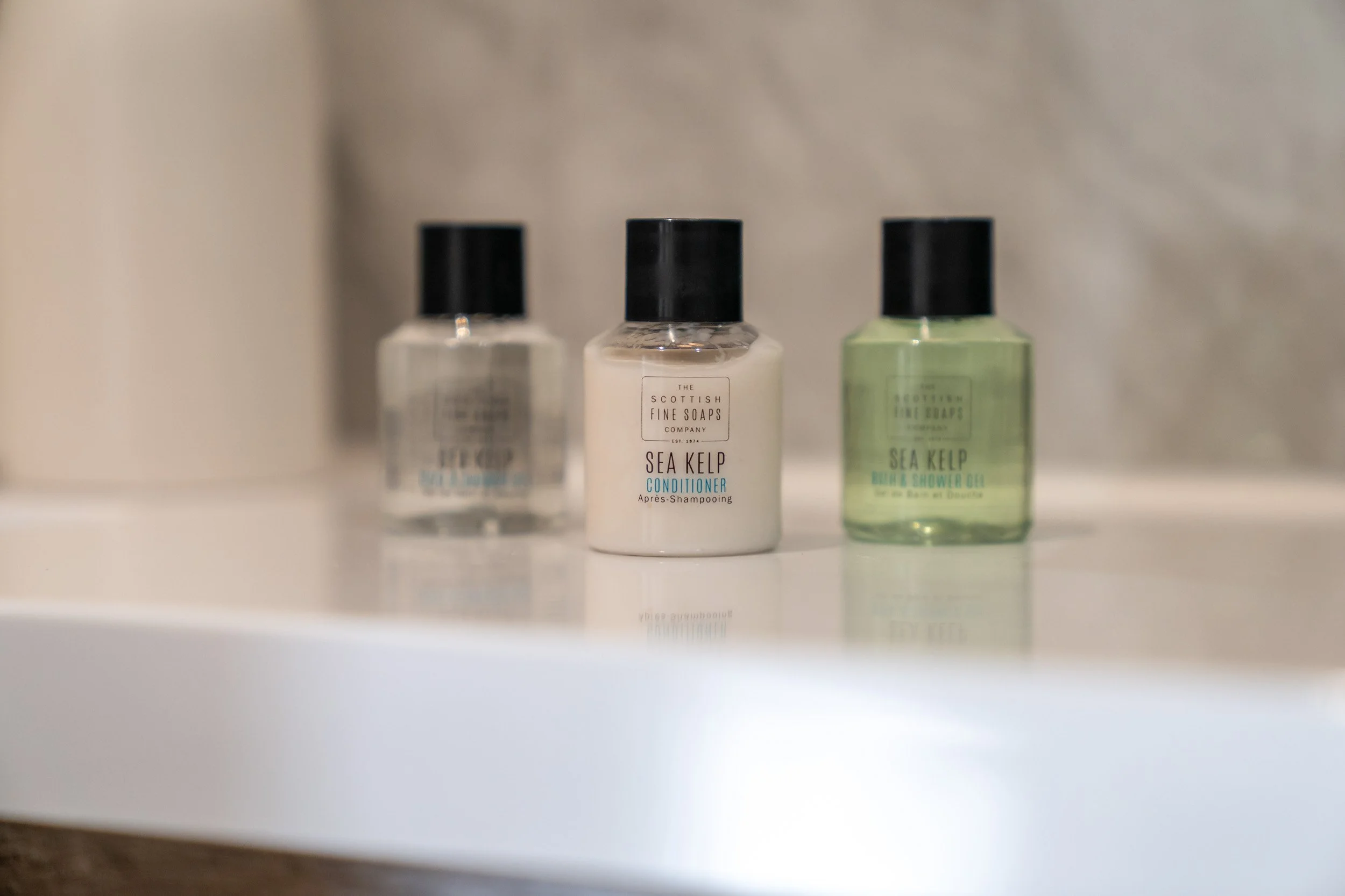 Three small bottles of toiletries on a bathroom shelf, with the central bottle labeled 'Sea Kelp Conditioner' and the other bottles being shampoo and shower oil.