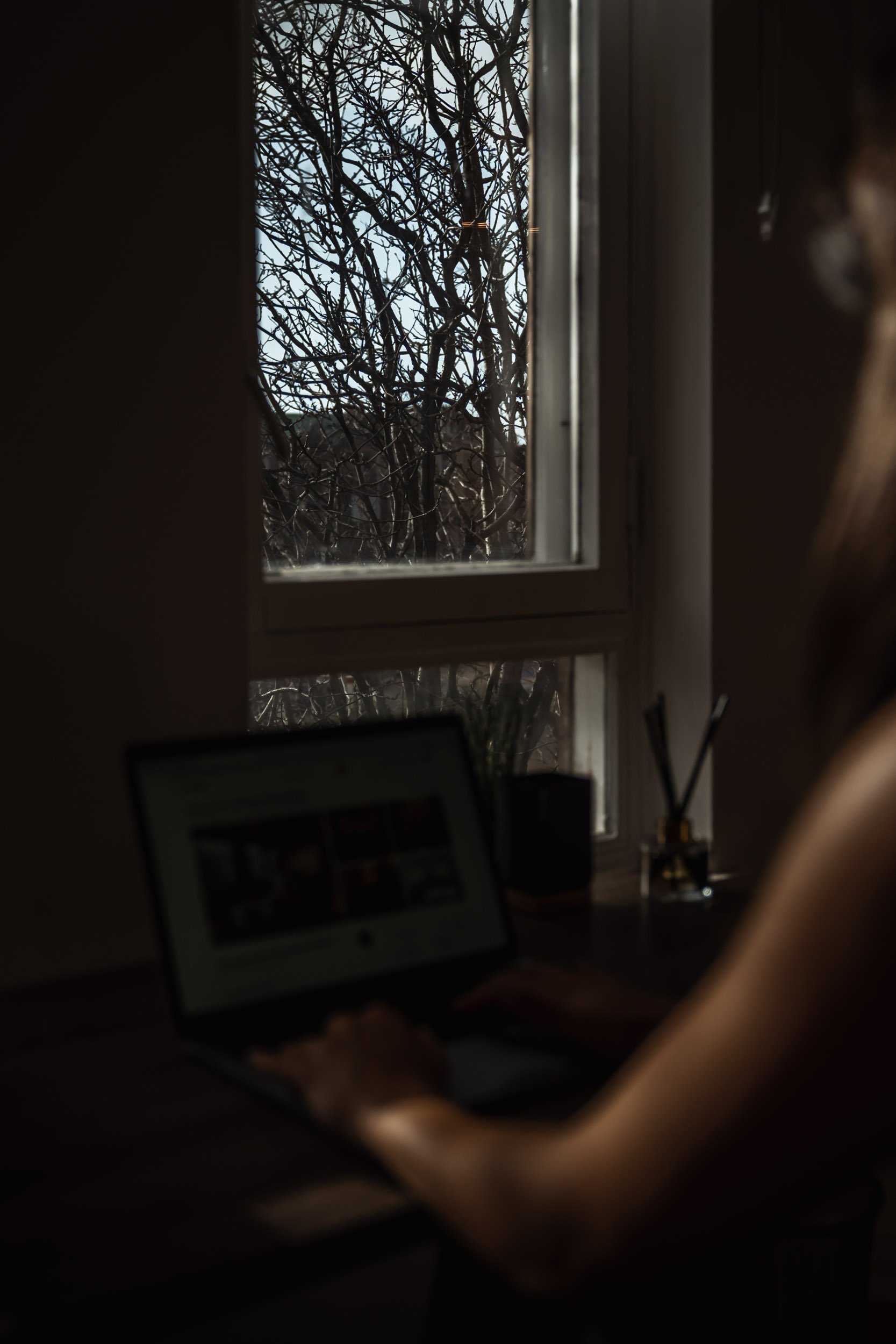 A person works on a laptop at a desk near a window with a view of a tree outside, during daylight.