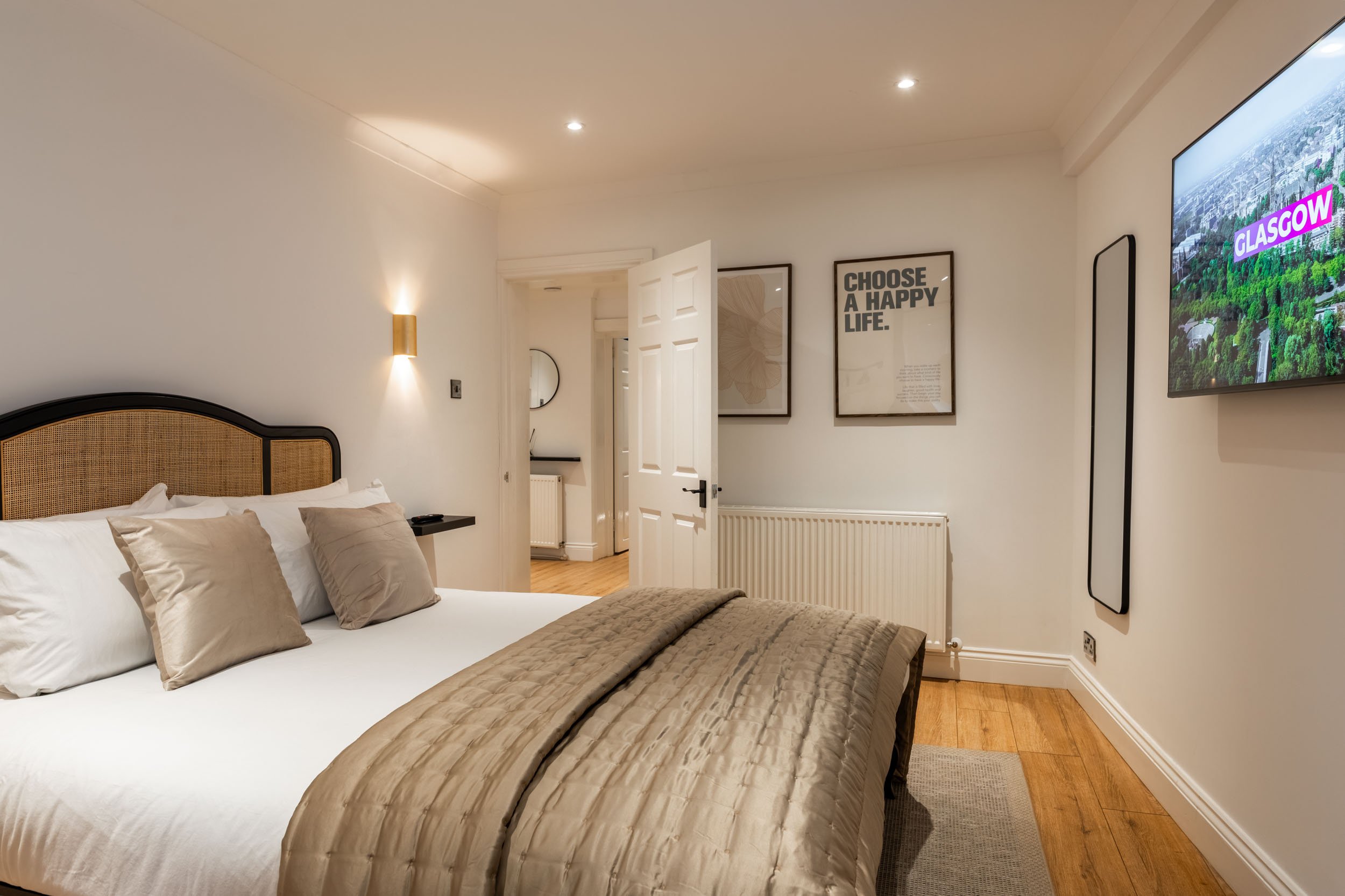 A neatly made bedroom with a large bed covered in white and beige linens and pillows. There are two framed posters on the wall, one says "Choose a Happy Life." and the other says "Glasgow" on a TV screen. The room has wood flooring, a door open to a 