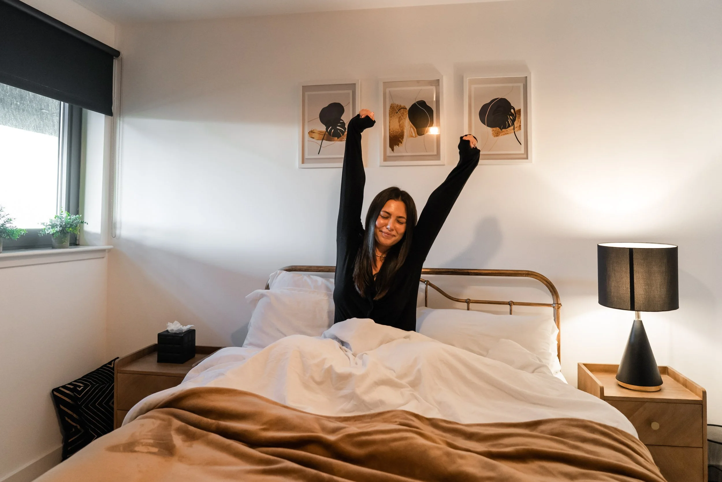 A woman waking up in bed stretching with arms raised, smiling, in a cozy bedroom with white walls, framed art above the bed, a bedside table with a black lamp, a tissue box, and a window with a partially drawn black blind and potted plants outside.