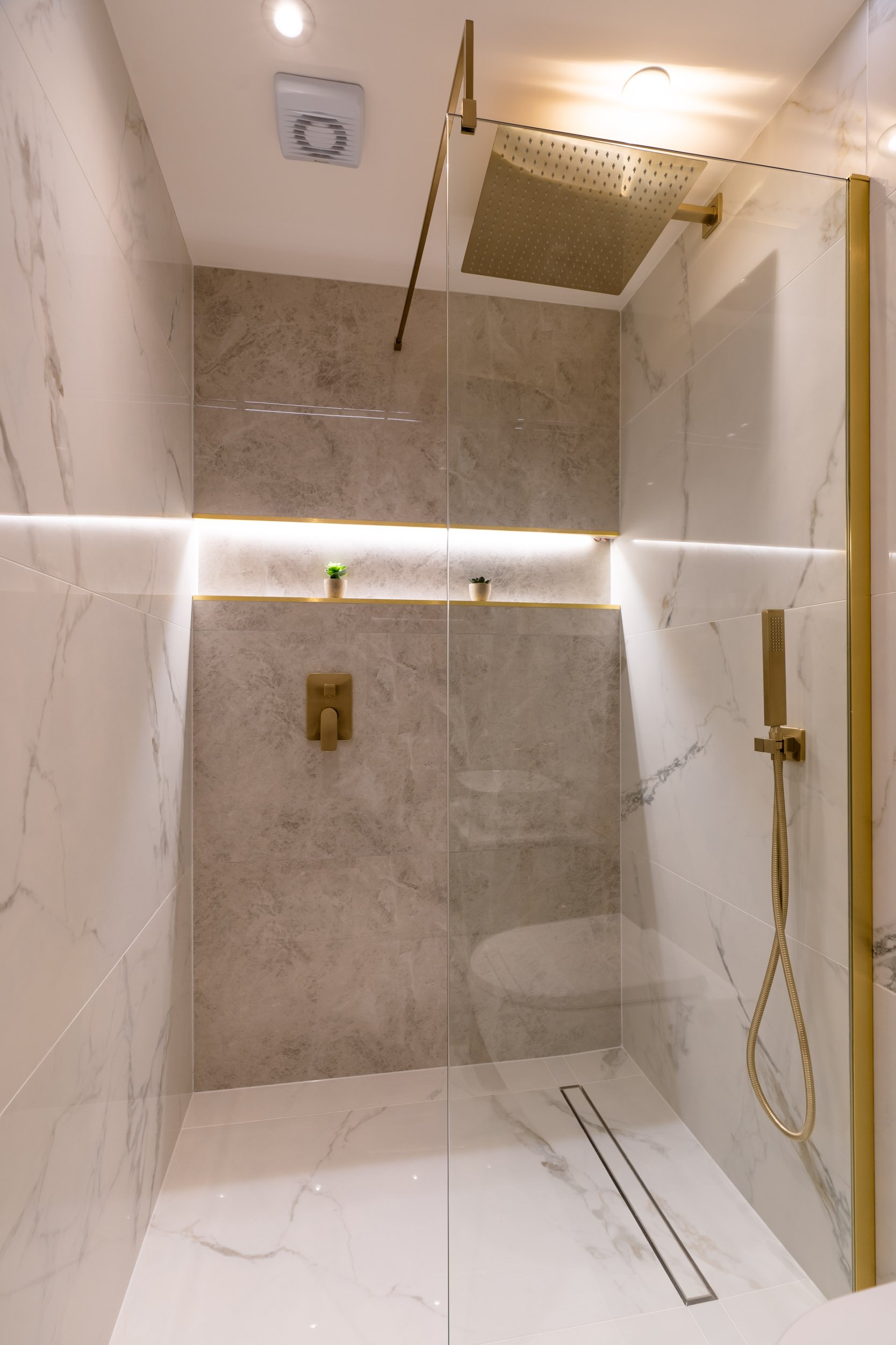 Modern bathroom shower area with marble tiles, a ceiling rain shower, handheld shower, small potted plants on a ledge, and warm lighting.